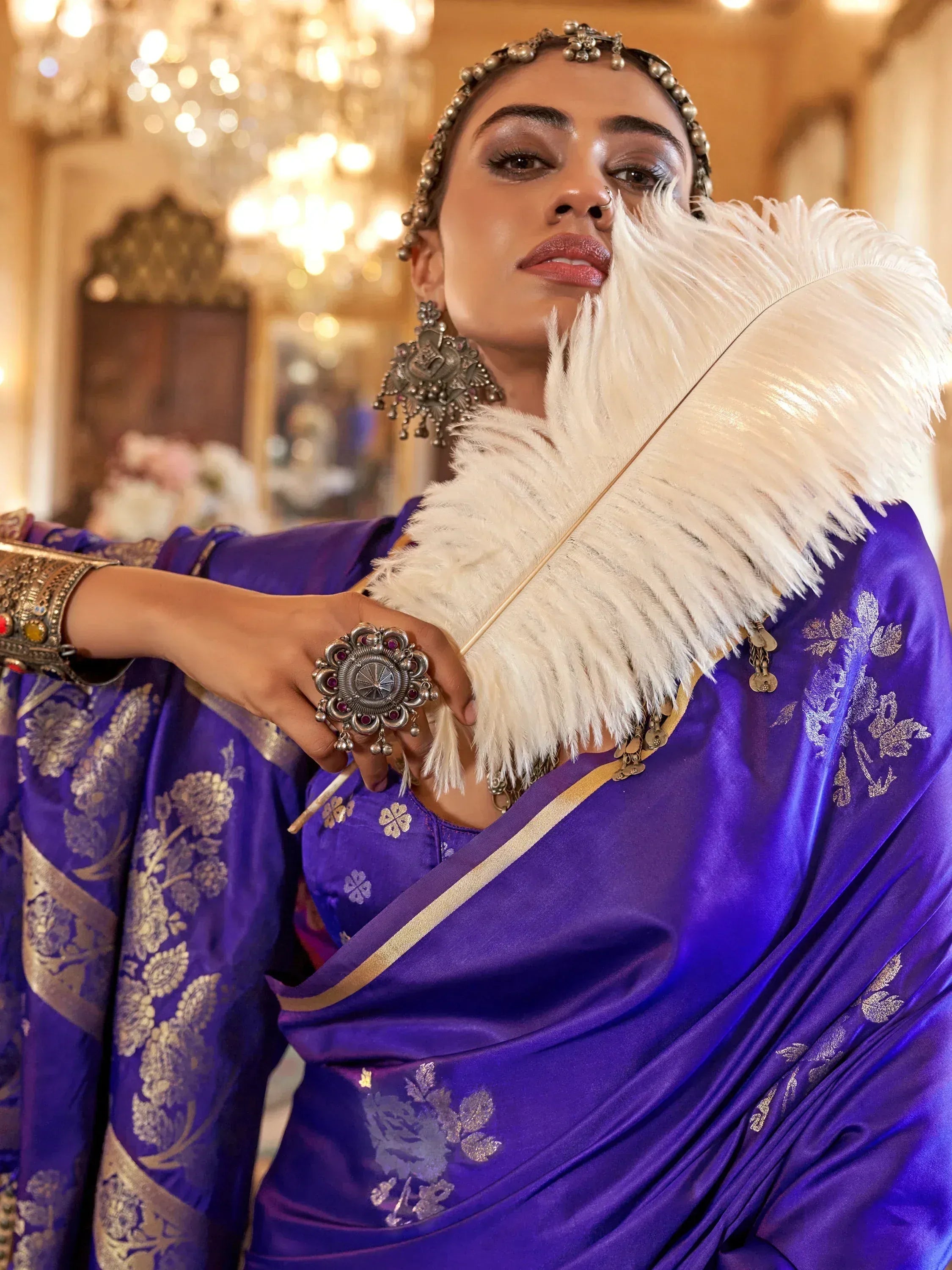 Elegant woman in royal purple ethnic saree with gold floral print, holding white feather fan, adorned with traditional jewelry.