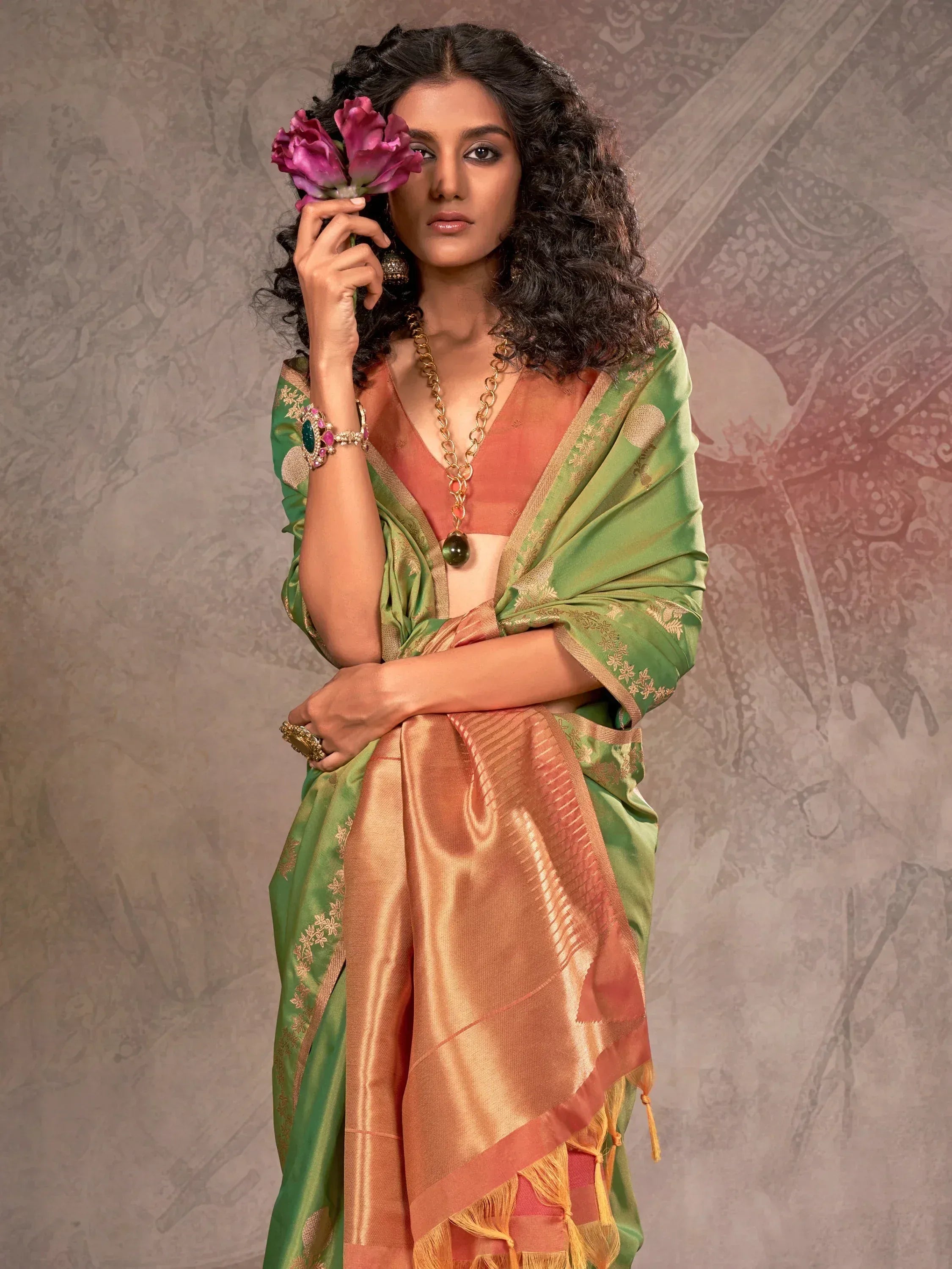 Woman in green and peach designer saree with gold embroidery, holding pink flowers, ethnic Indian wear