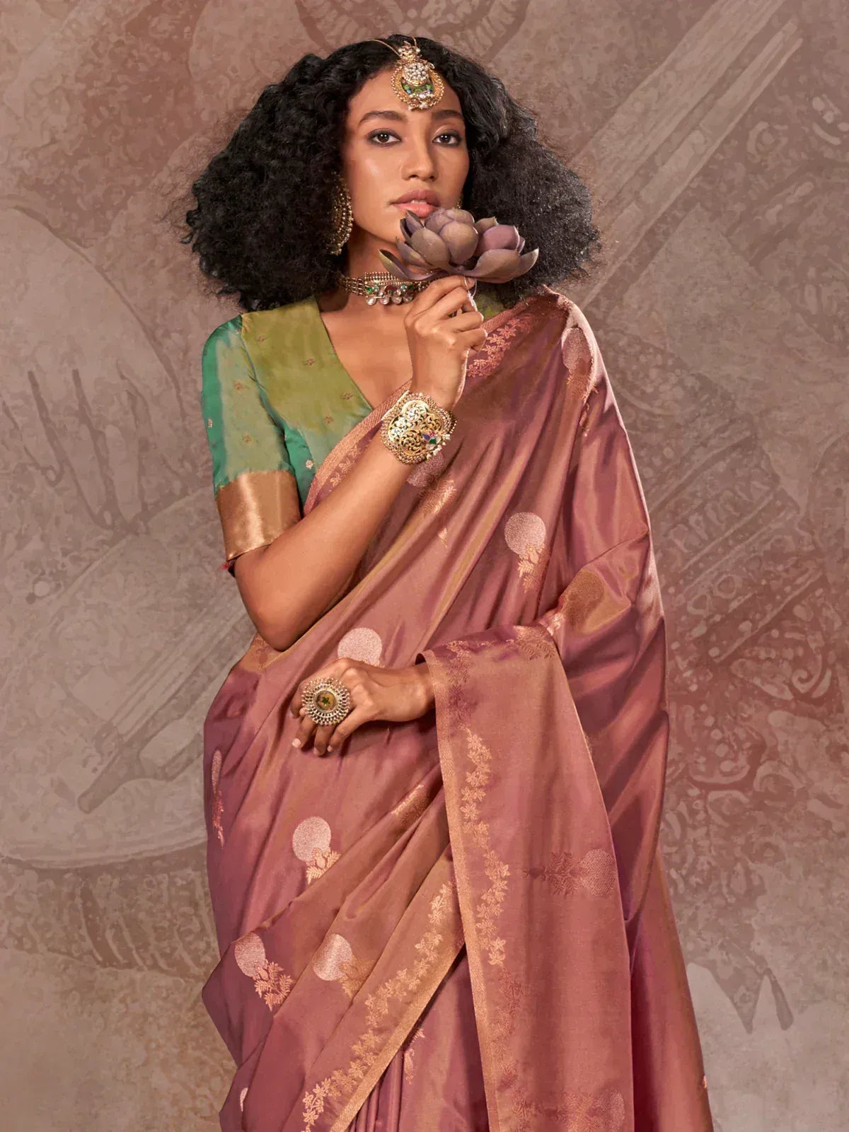 Woman in elegant mauve silk saree with gold motifs, green blouse, and traditional jewelry holding lotus flower