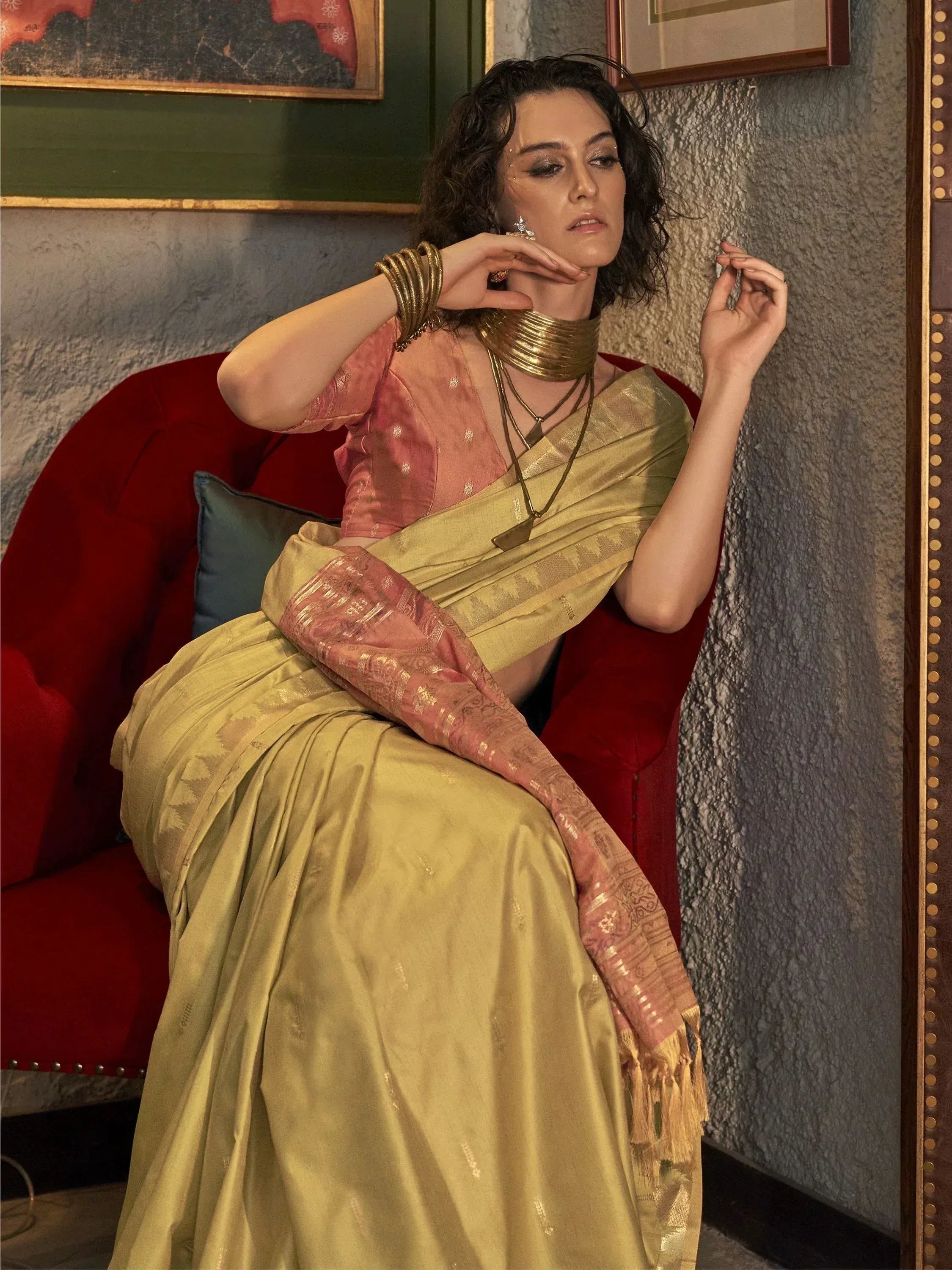 Woman in elegant gold and pink saree with traditional jewelry, seated on red velvet chair