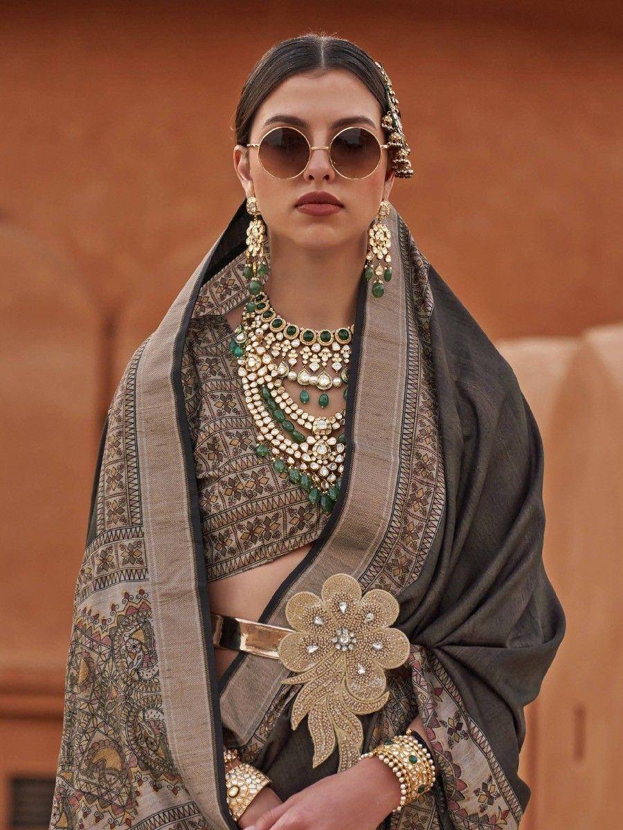 Woman in ethnic Indian saree with intricate gold and green jewelry, styled with sunglasses and floral belt.