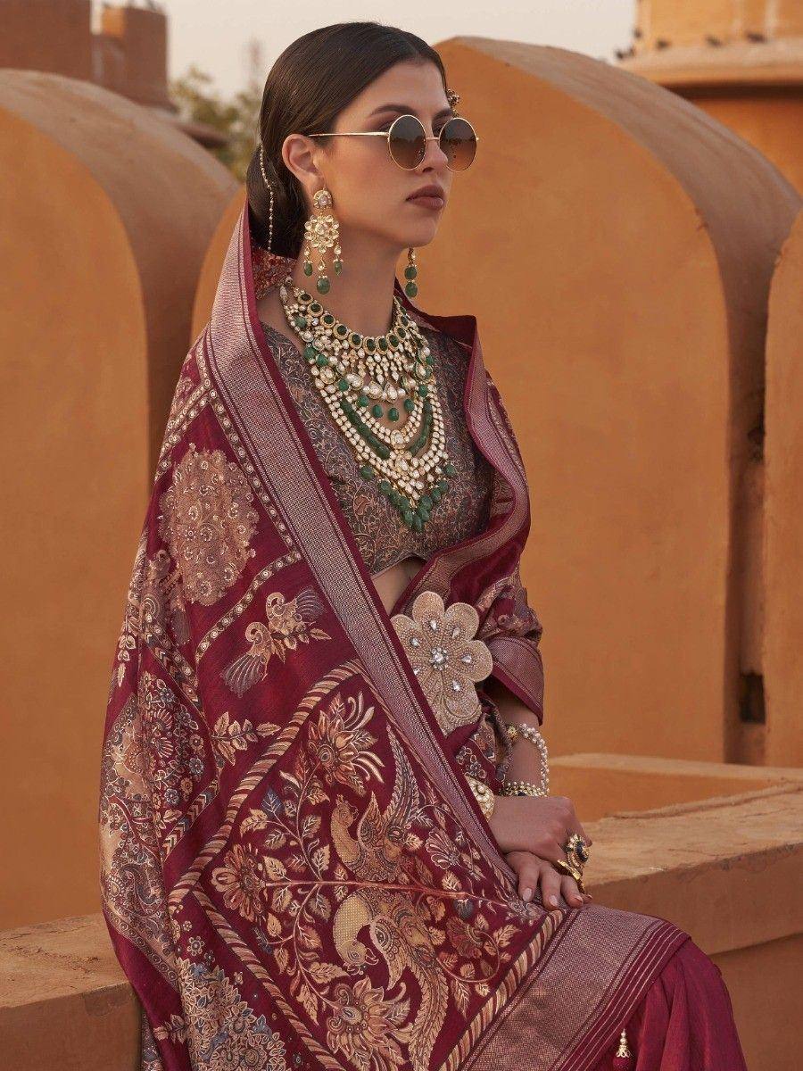 Woman in maroon embroidered saree with gold peacock motifs, green jewelry, and round sunglasses