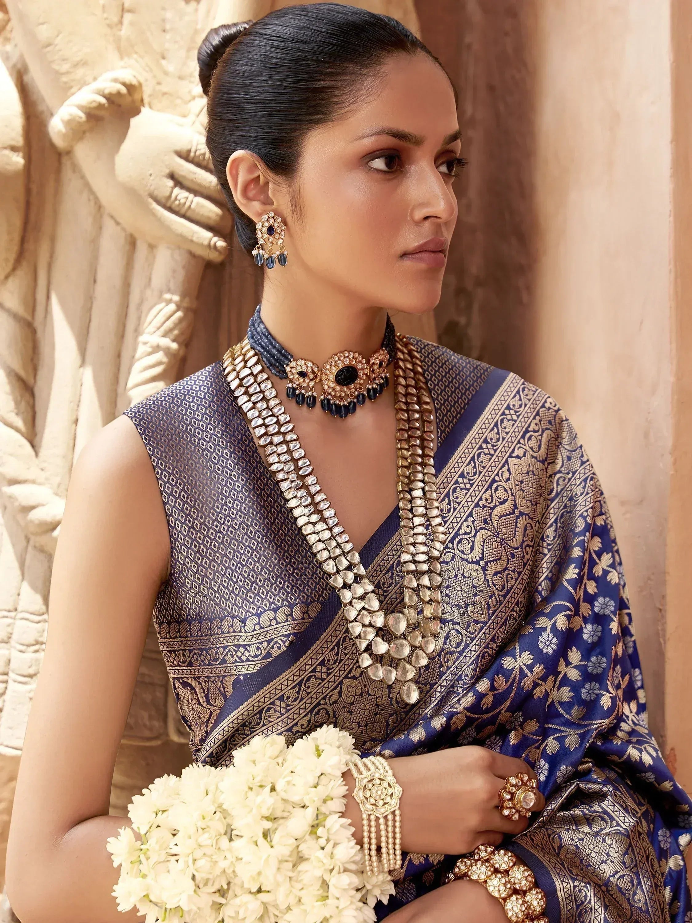 Elegant woman in blue and gold designer saree with traditional jewelry, holding white flowers