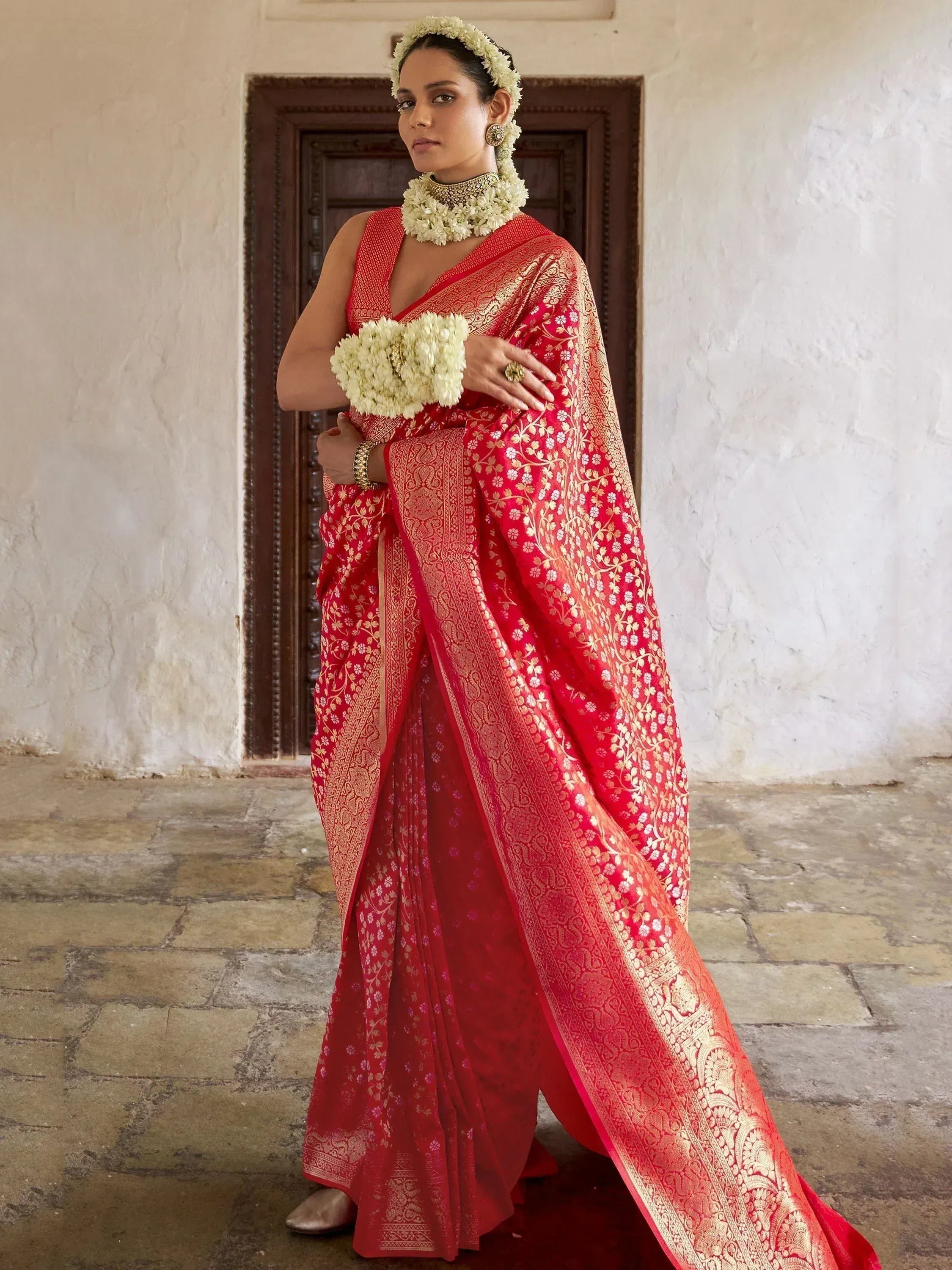 Red and gold designer saree with floral motifs, paired with traditional jewelry and floral accessories