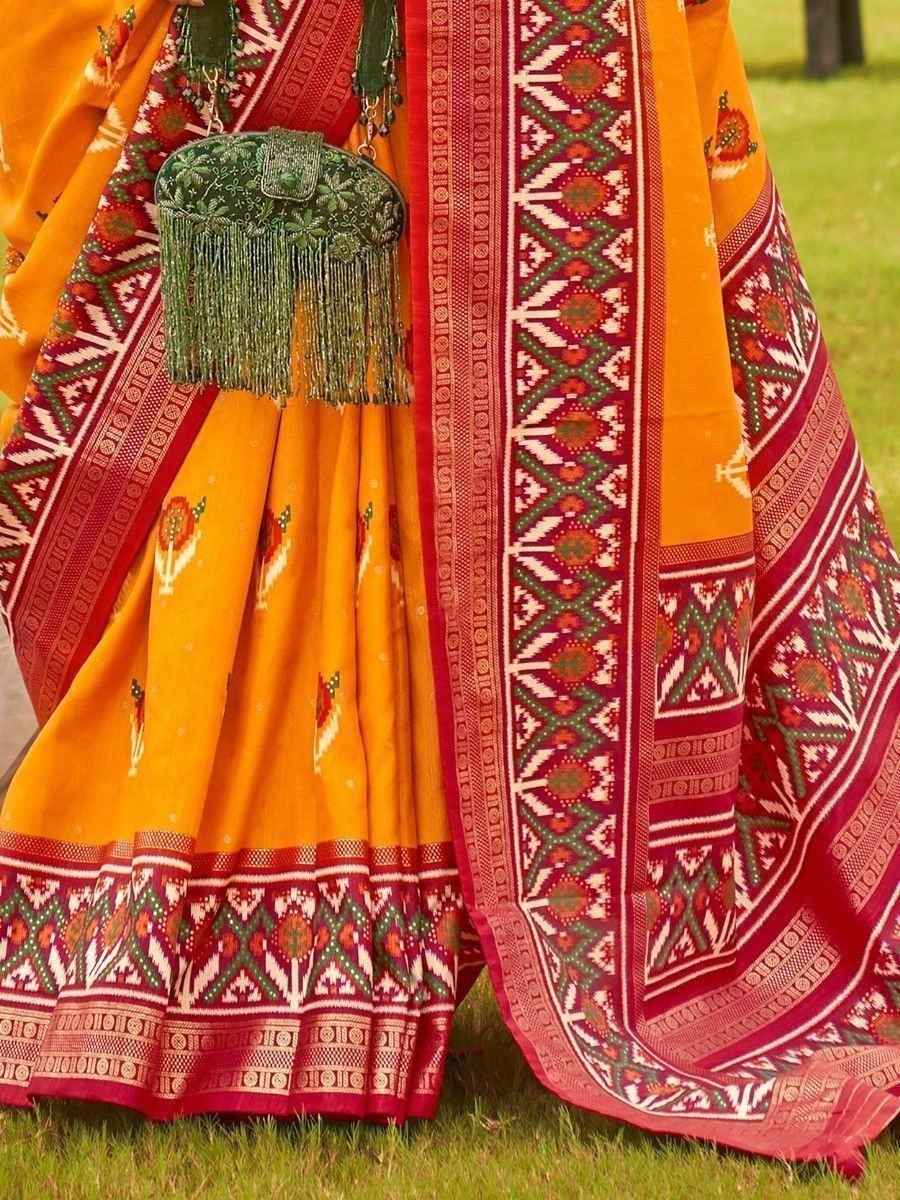 Vibrant orange and red ethnic saree with intricate geometric patterns and green tassel bag, perfect for festive occasions.