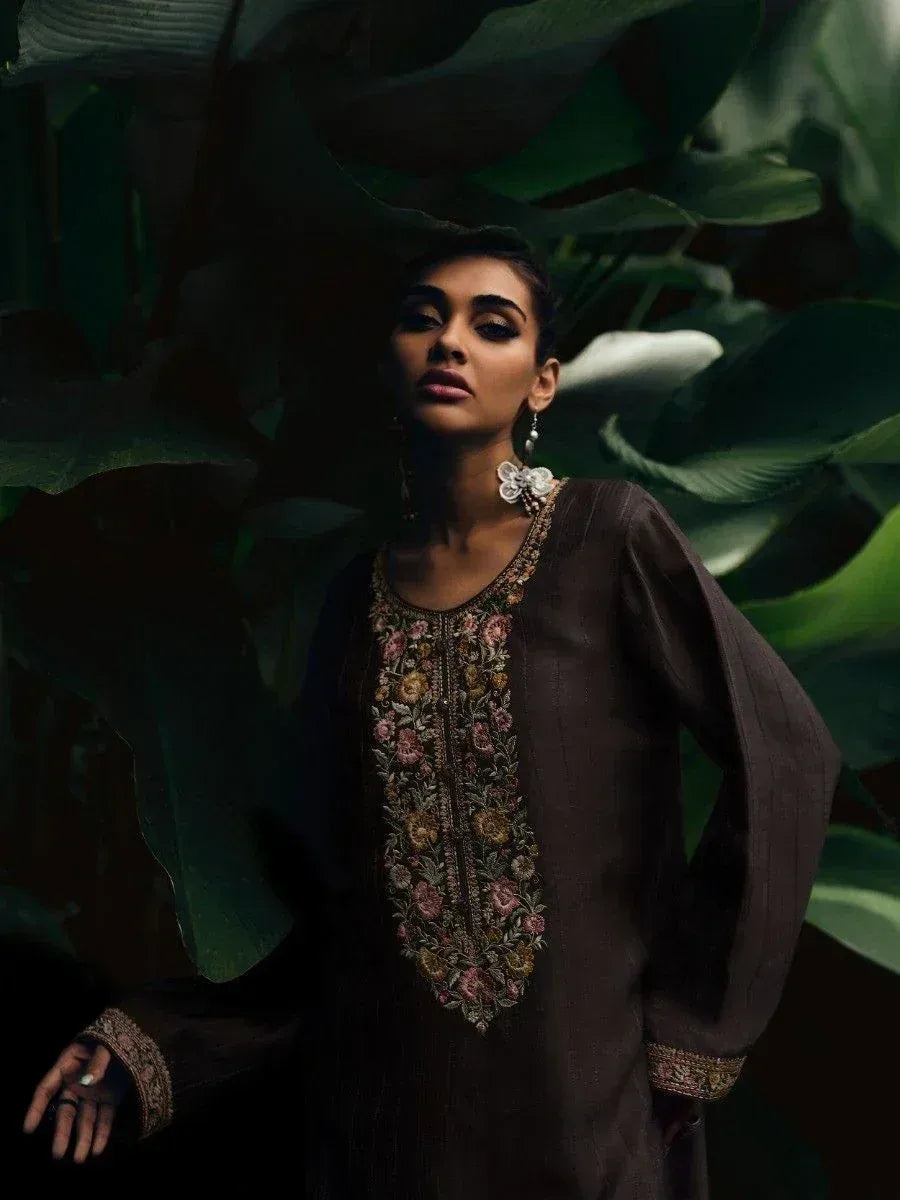 Woman in brown embroidered ethnic kurta with floral detailing, styled with statement earrings against lush green foliage.