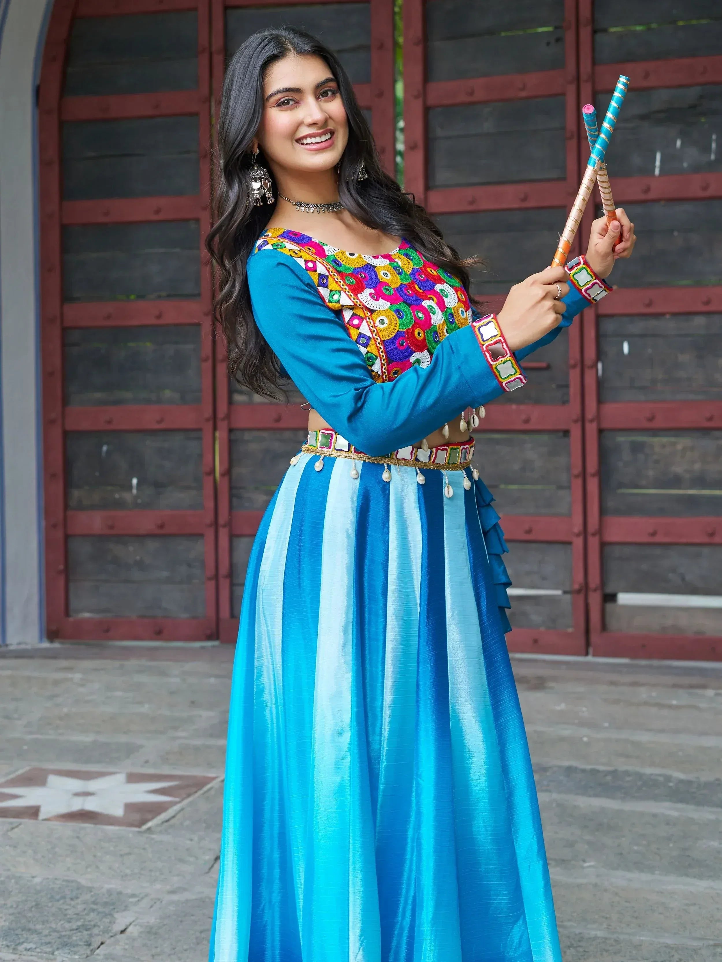 Woman in vibrant blue embroidered lehenga choli with mirror work, holding dandiya sticks for festive dance