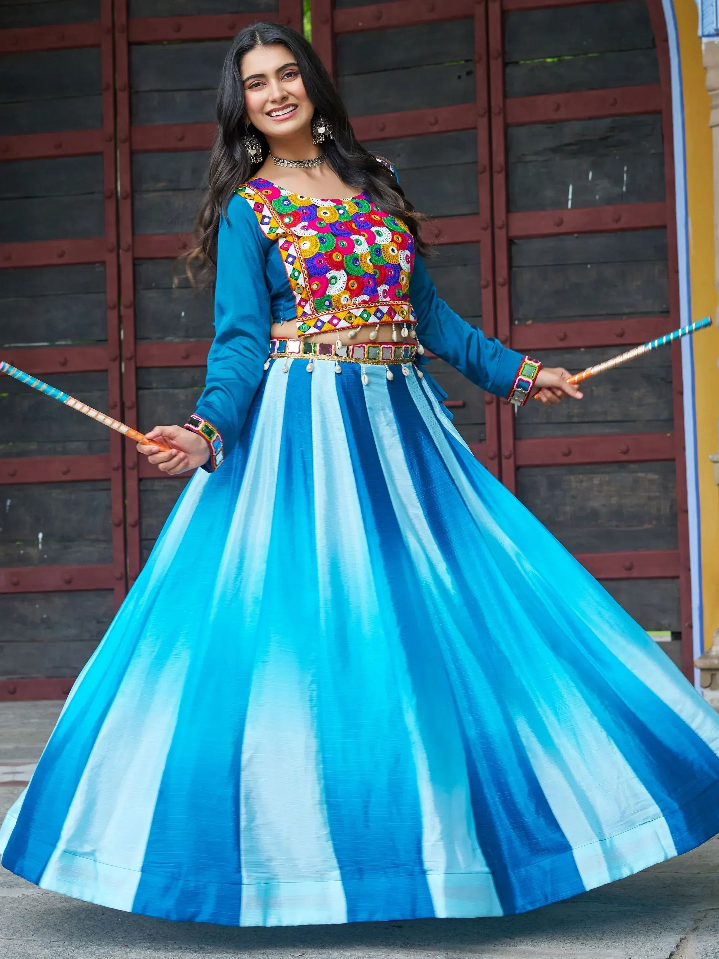 Woman in vibrant blue striped lehenga choli with mirror work, holding dandiya sticks for festive Indian dance