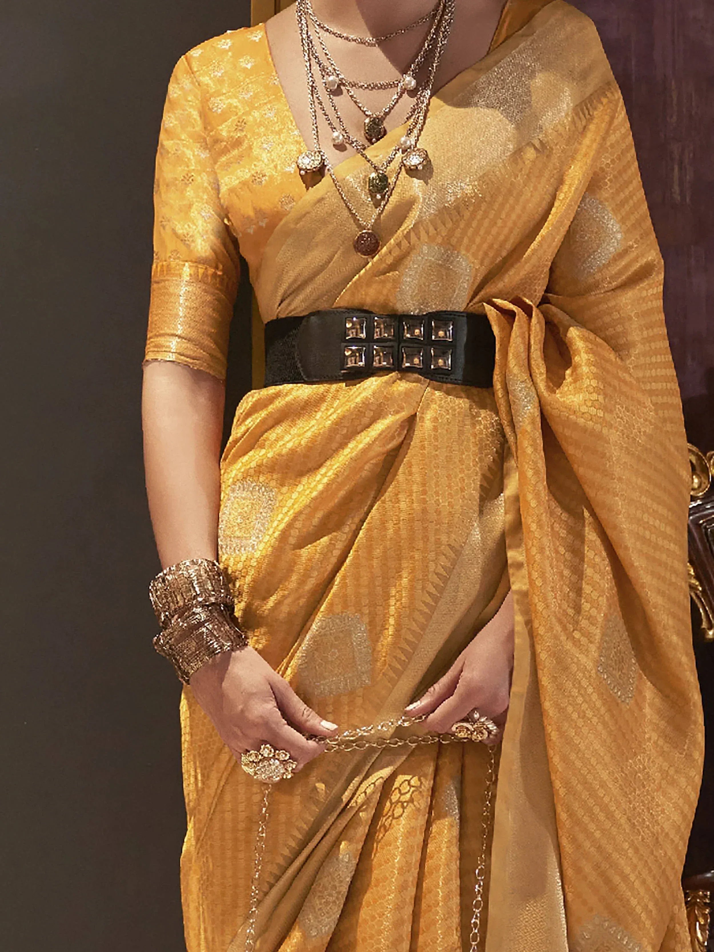 Elegant golden saree with intricate patterns, styled with layered necklaces and a black studded belt for festive occasions.