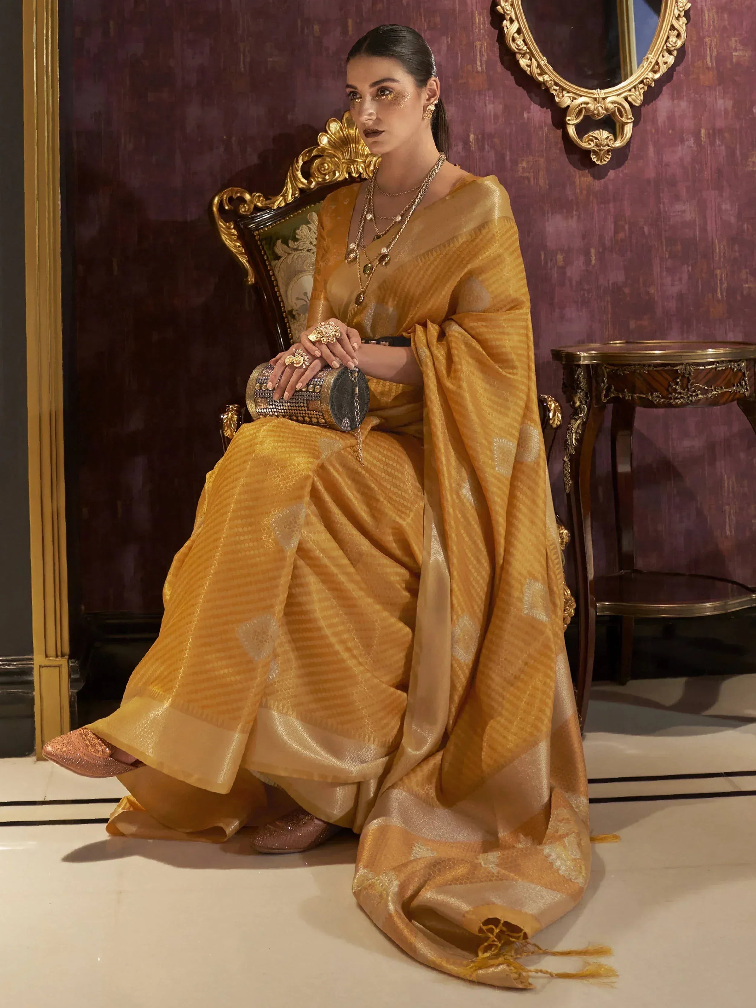 Elegant woman in golden designer saree with intricate patterns, seated on ornate chair, perfect for ethnic occasions