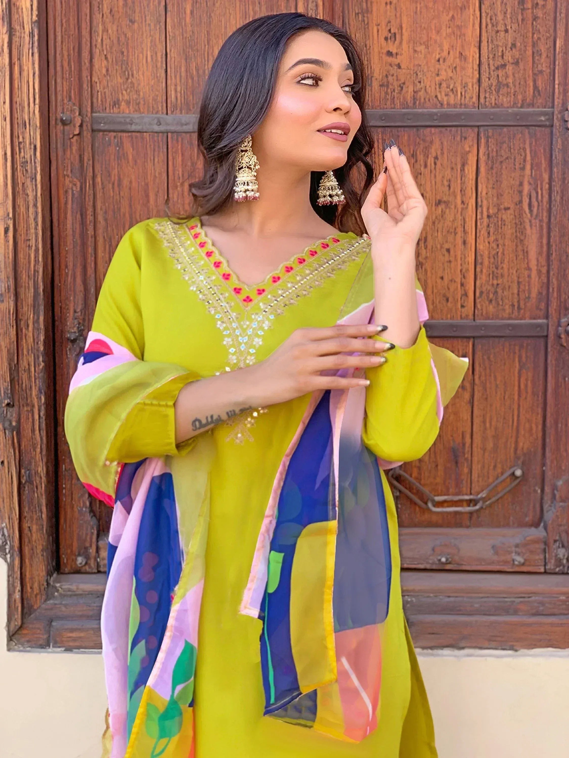 Woman in vibrant green embroidered salwar kameez with colorful dupatta, standing by wooden door