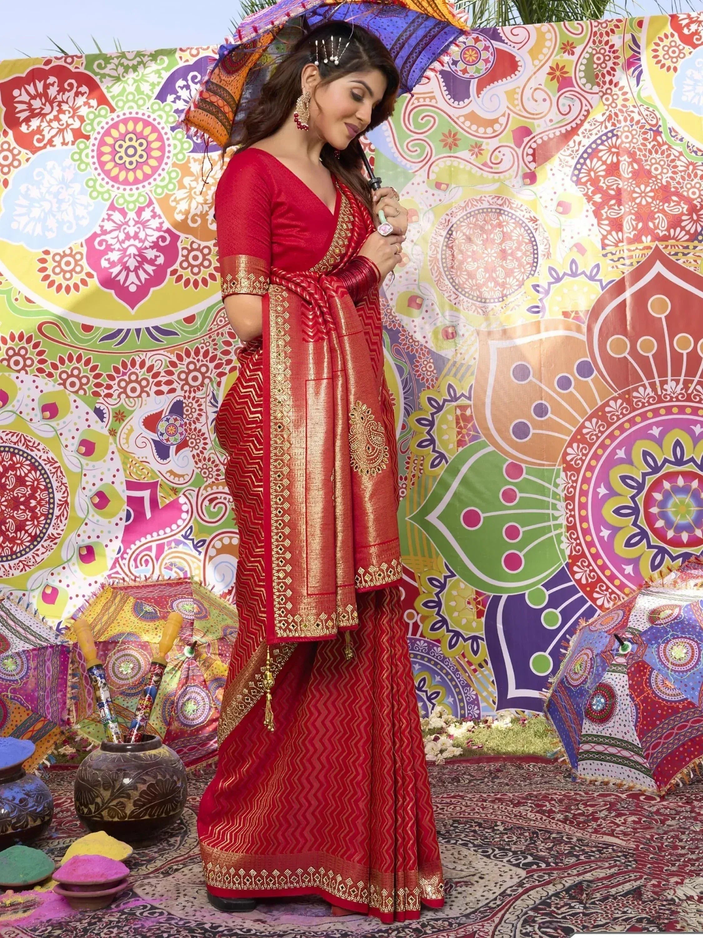Woman in red ethnic saree with gold embroidery, holding colorful umbrella against vibrant patterned backdrop