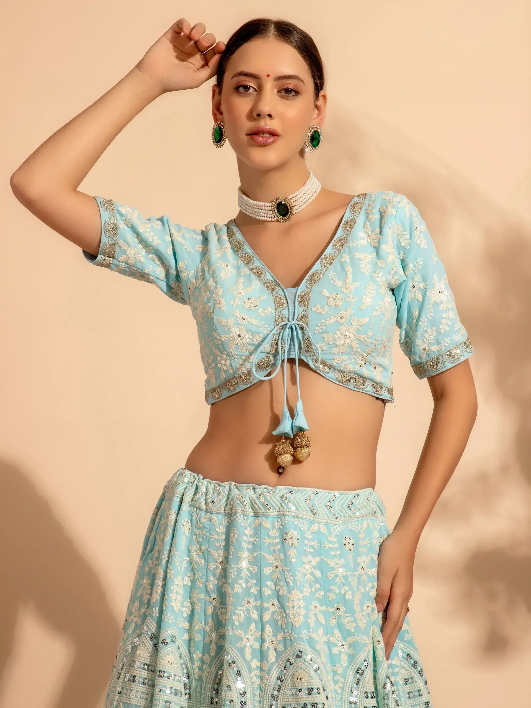 Light blue embroidered lehenga choli with floral motifs, gold detailing, and tassel tie, styled with green gem jewelry.