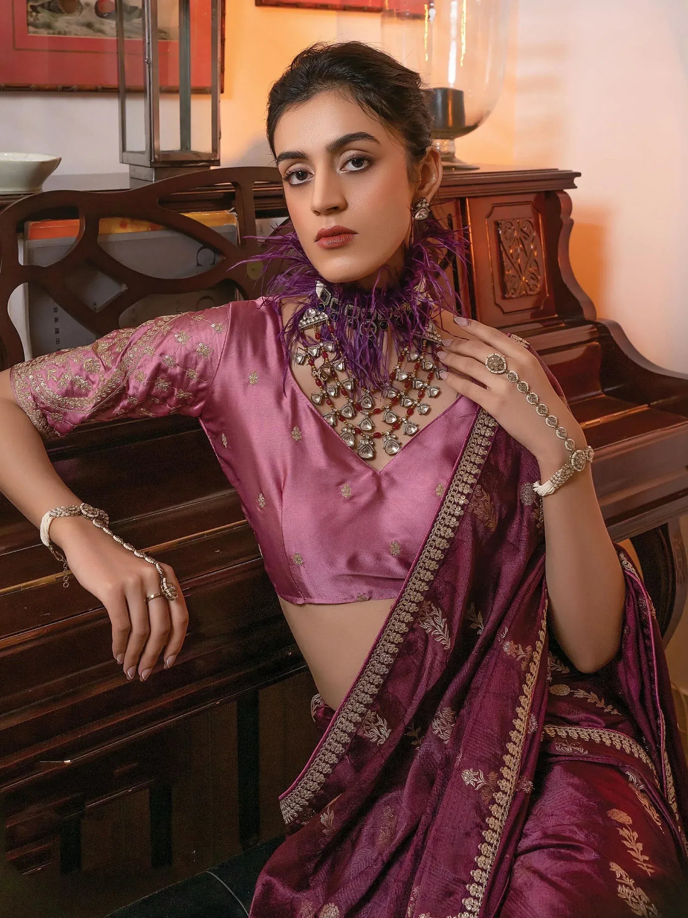 Elegant woman in purple silk saree with gold embroidery, styled with traditional jewelry and feathered neckpiece.