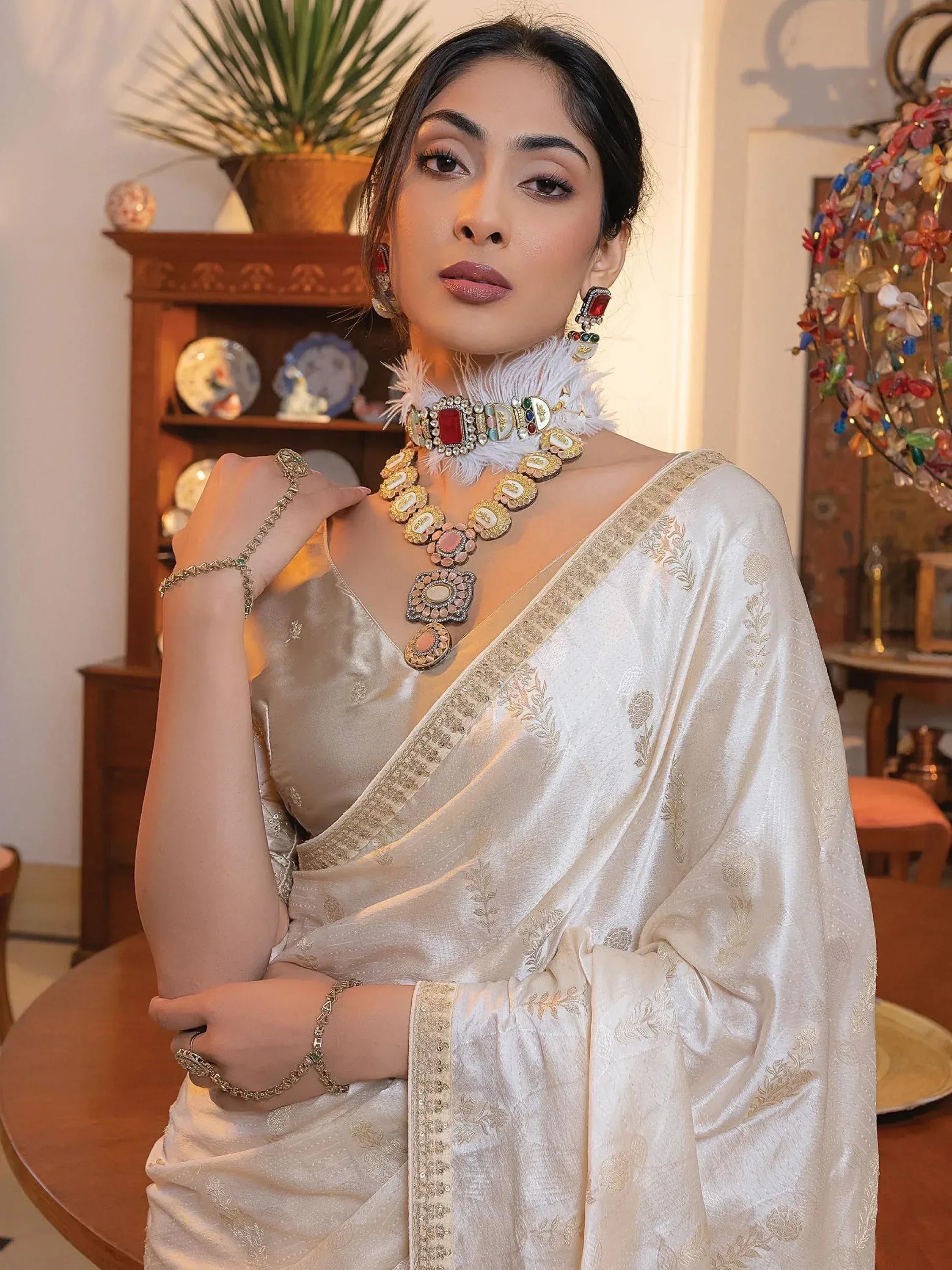 Elegant woman in white embroidered saree with gold detailing, wearing statement jewelry and feather choker.