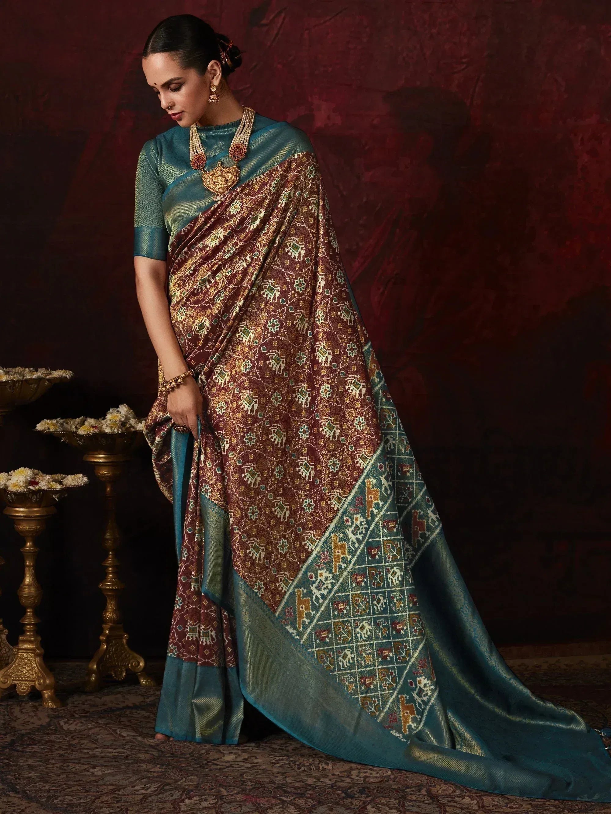 Woman in elegant teal and maroon ethnic saree with intricate elephant motifs, traditional jewelry, and floral decor.