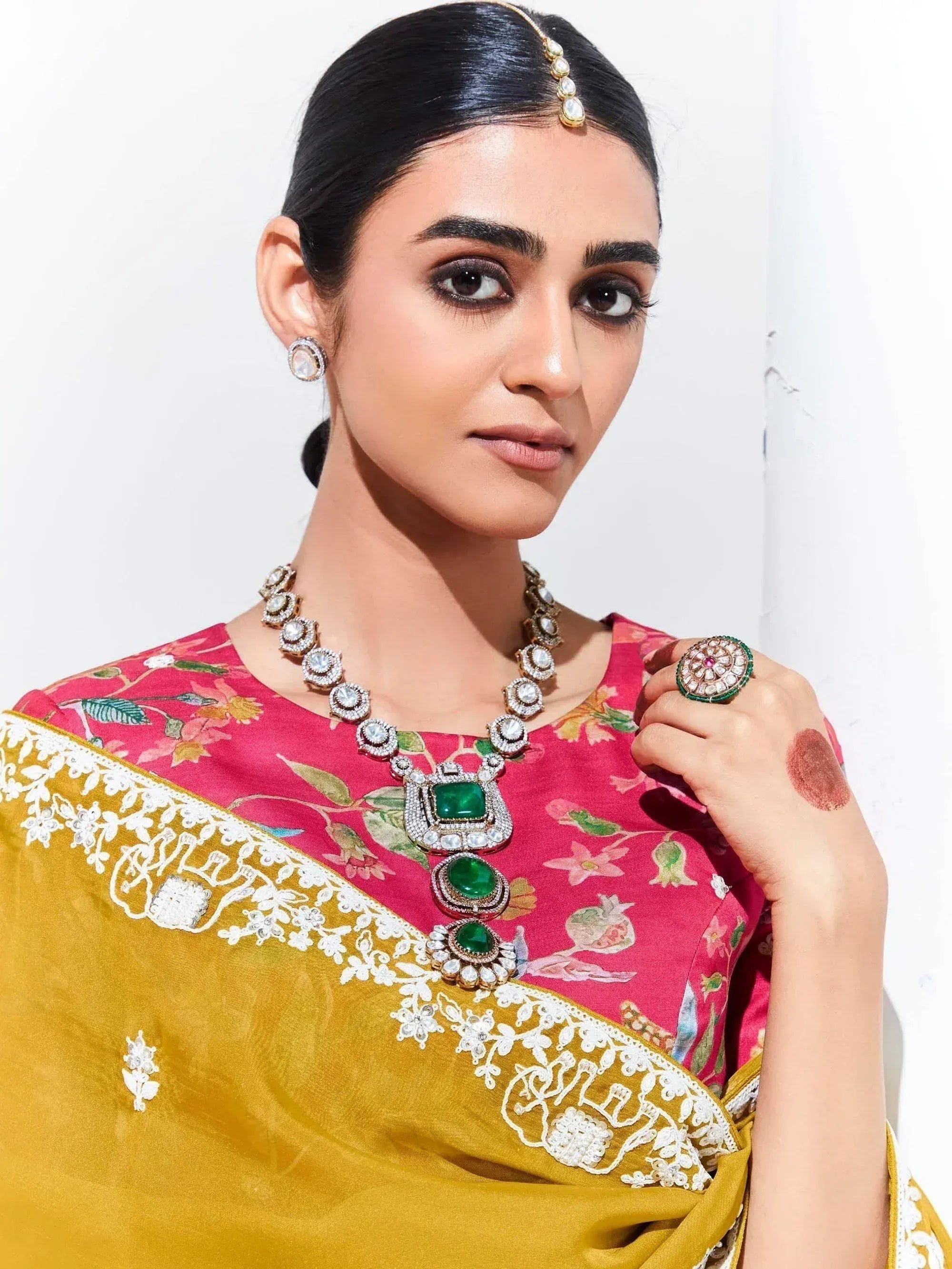 Woman in pink floral blouse & gold saree with emerald jewelry, perfect for Indian ethnic wear occasions.