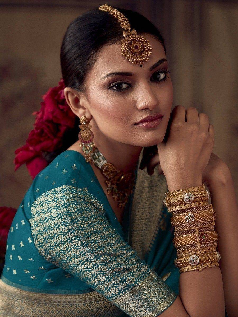 Woman in teal designer saree with gold embroidery, traditional Indian jewelry, and red floral hair accessory