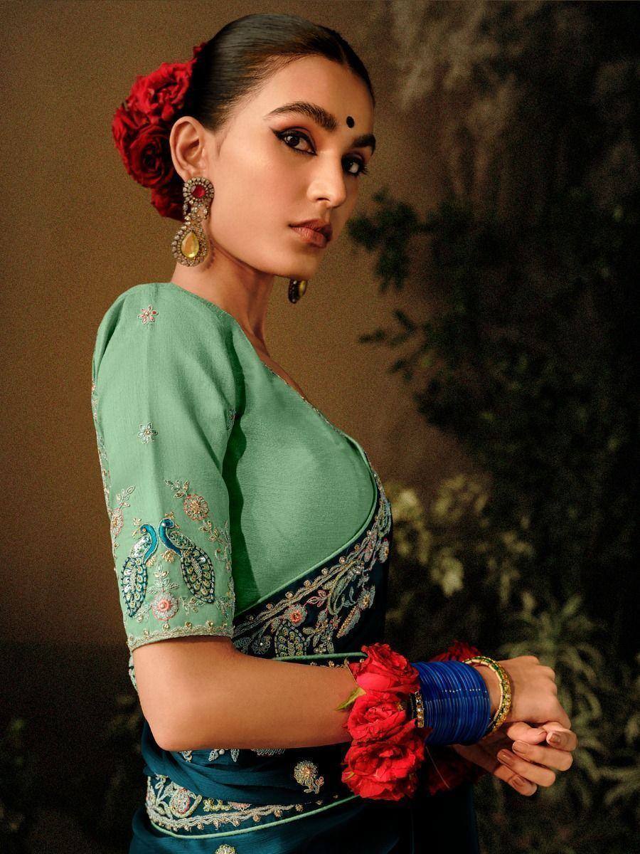 Elegant woman in teal embroidered saree with peacock motifs, red floral hair accessory, and blue bangles.