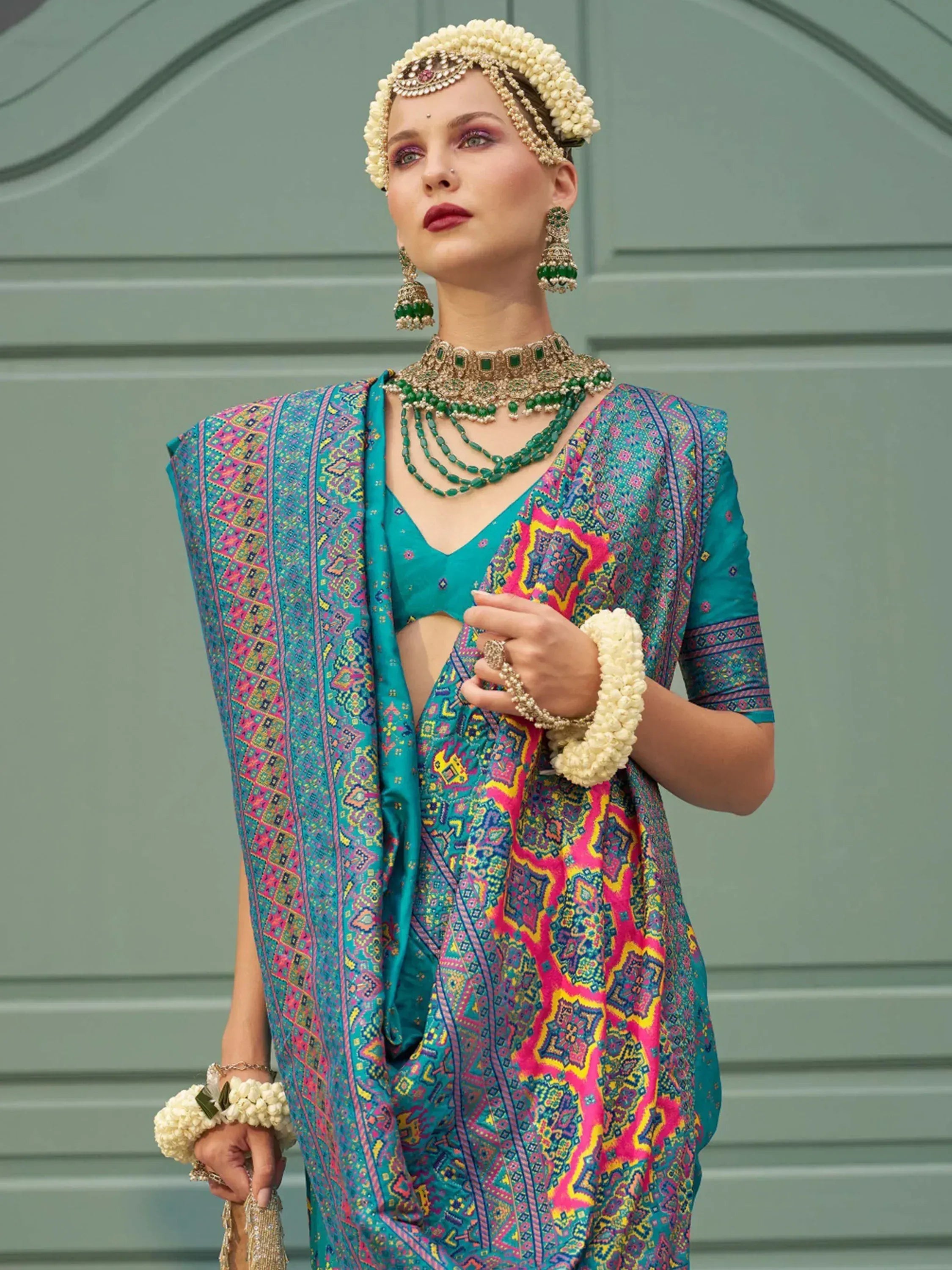 Woman in vibrant teal and pink ethnic saree with floral jewelry, perfect for Indian weddings or festive occasions.