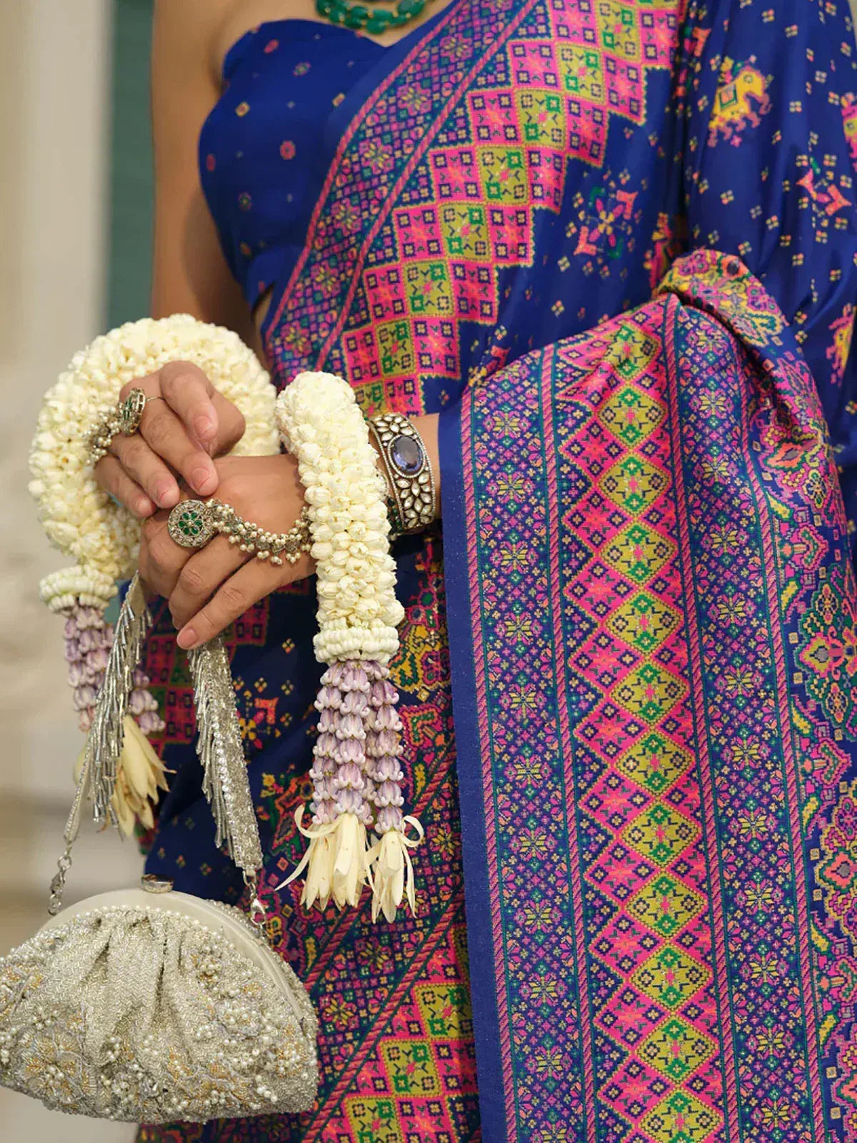 Vibrant blue saree with pink and green geometric print, paired with floral garlands and embellished clutch for ethnic wear.