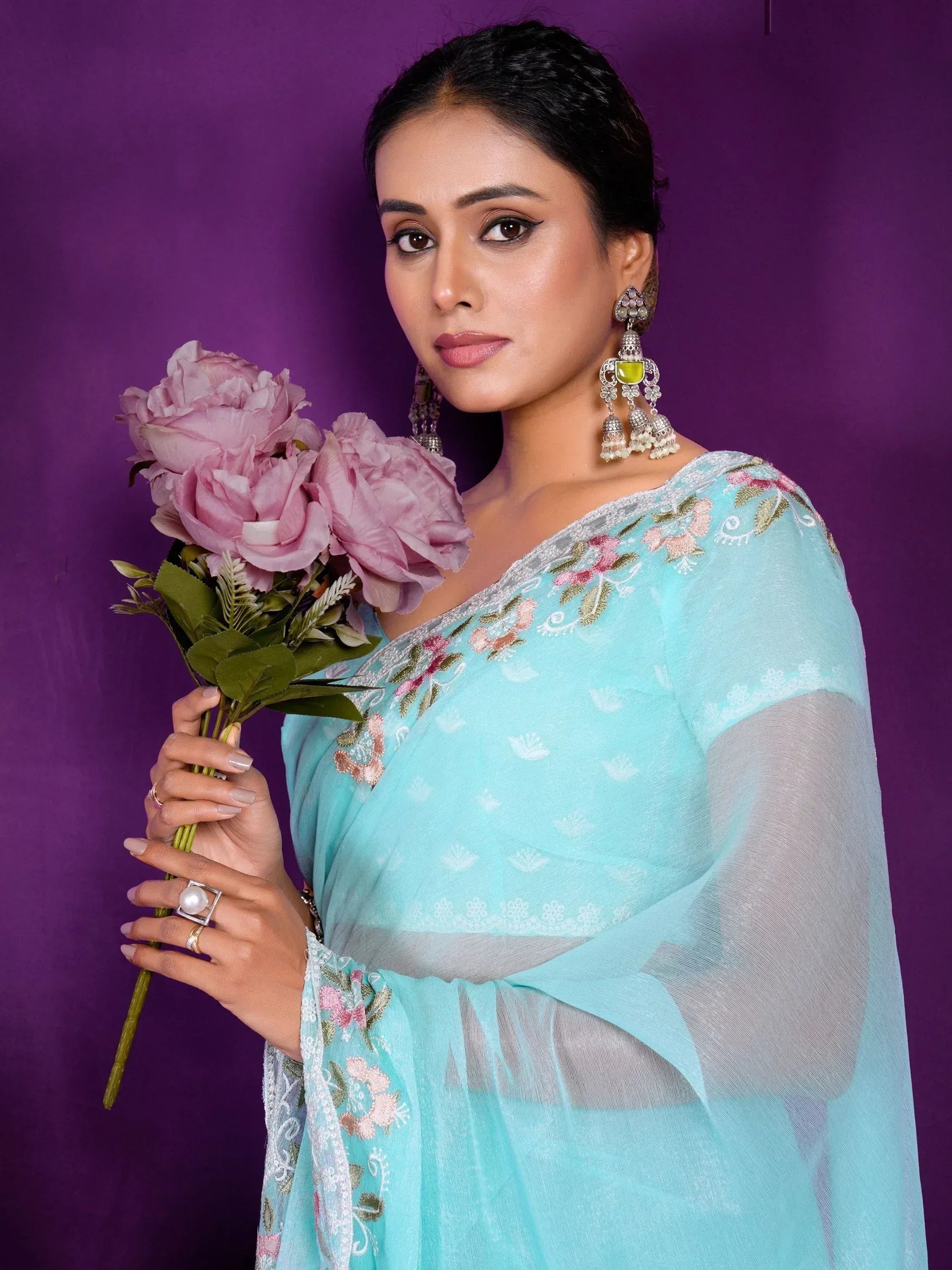 Woman in light blue embroidered ethnic gown with floral details, holding pink roses against purple background