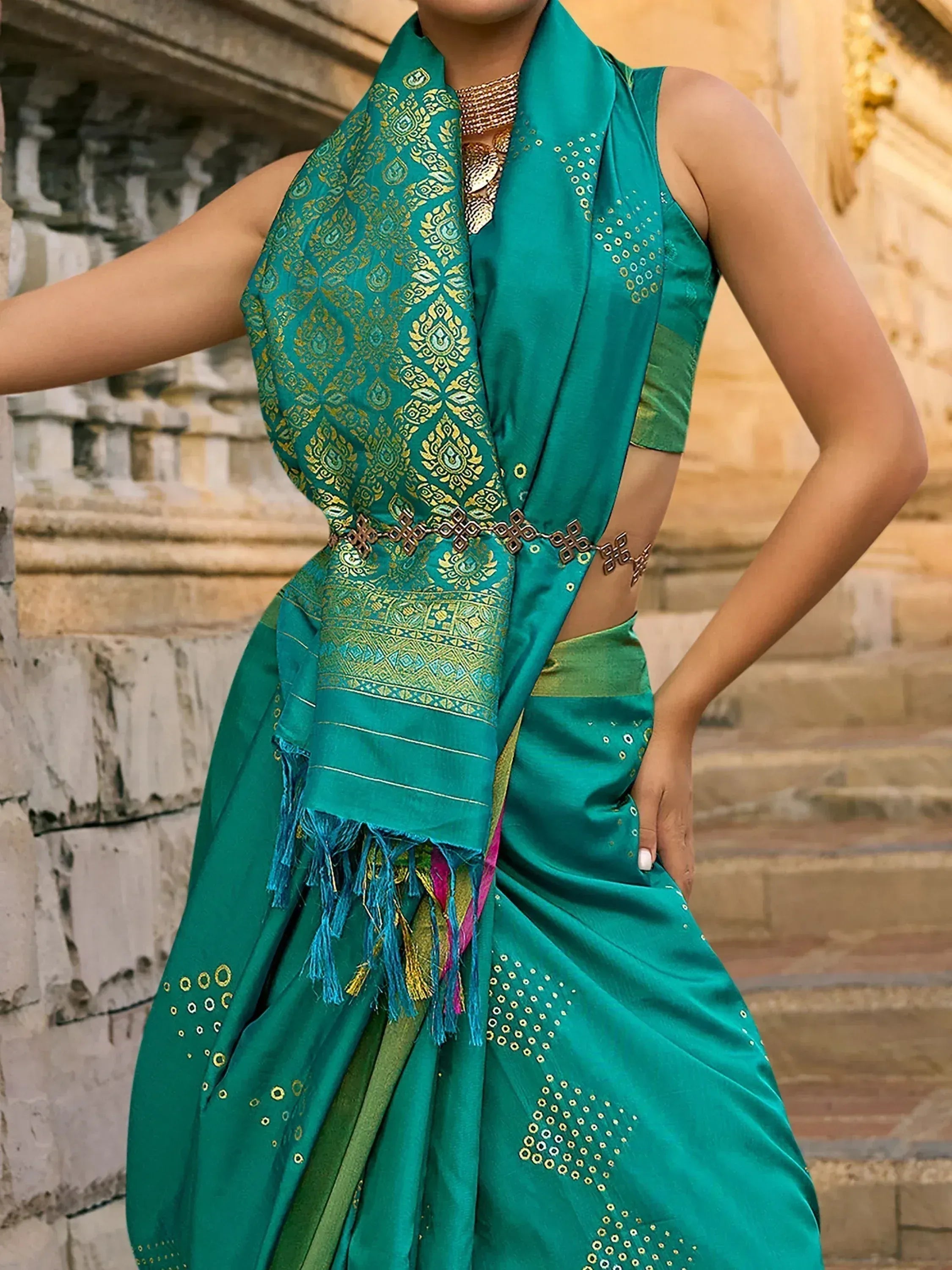 Teal green designer saree with gold embroidery and mirror work, paired with sleeveless blouse and temple backdrop