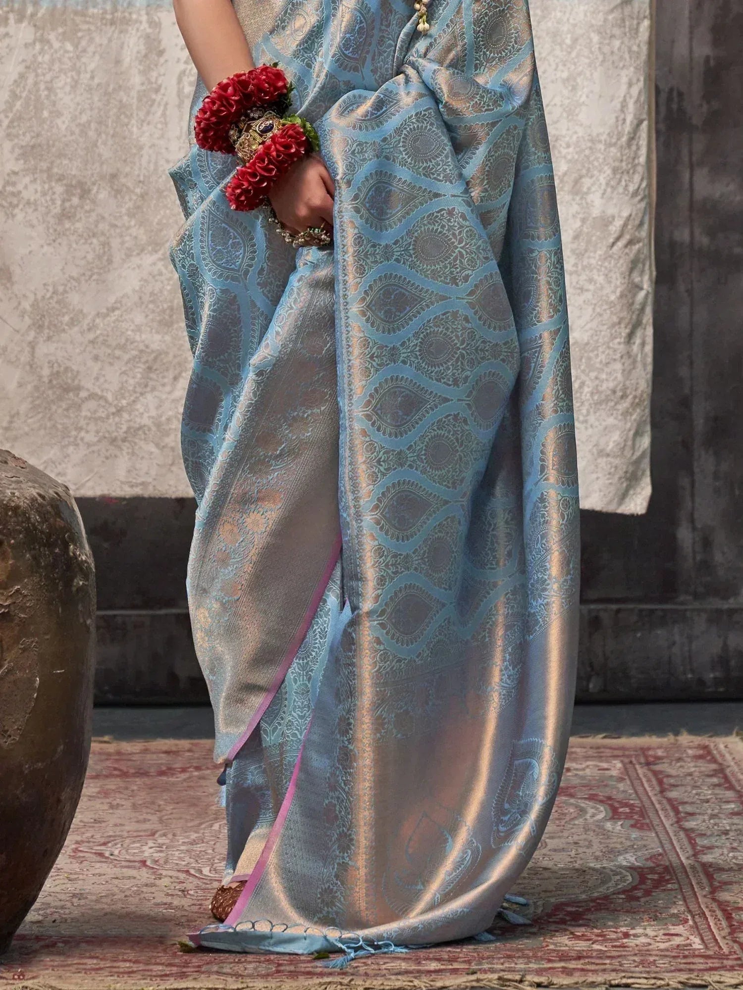 Light blue designer saree with intricate paisley weave and pink border, styled with floral wrist accessory