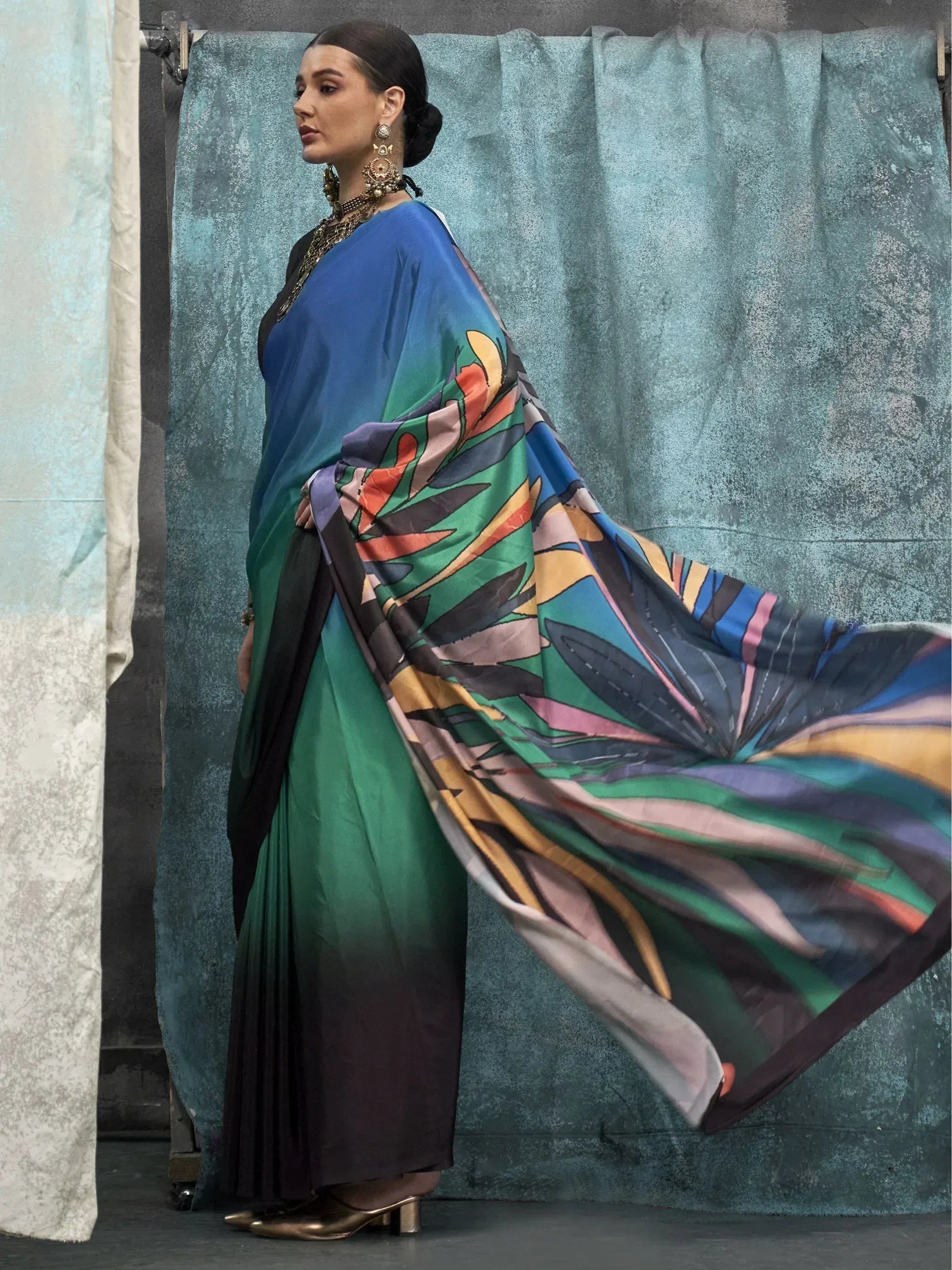 Woman in vibrant printed saree with ombre green-blue drape, ethnic jewelry, and gold heels