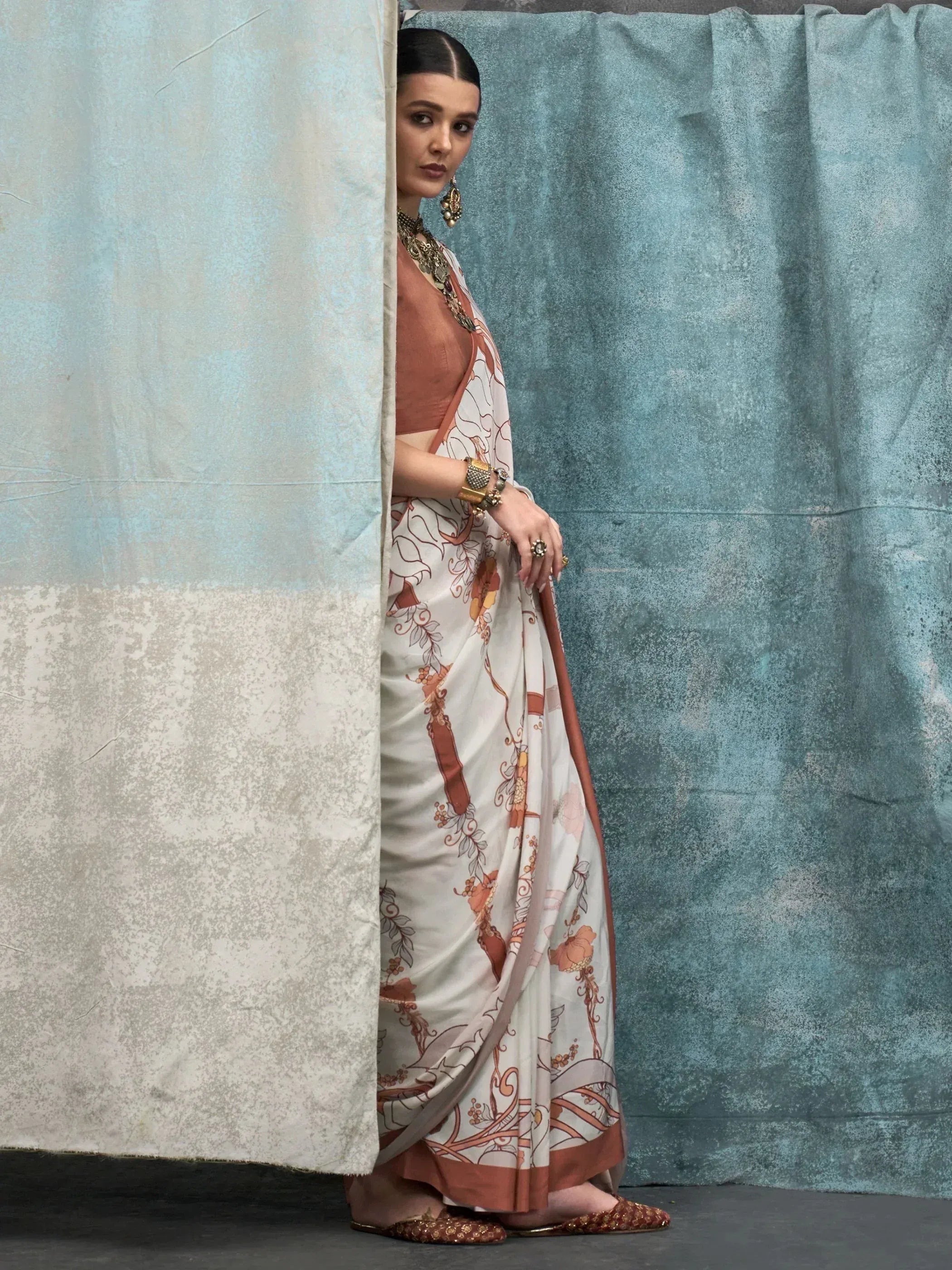 Woman in elegant floral saree with brown blouse, traditional jewelry, and ethnic flats, peeking from behind curtain