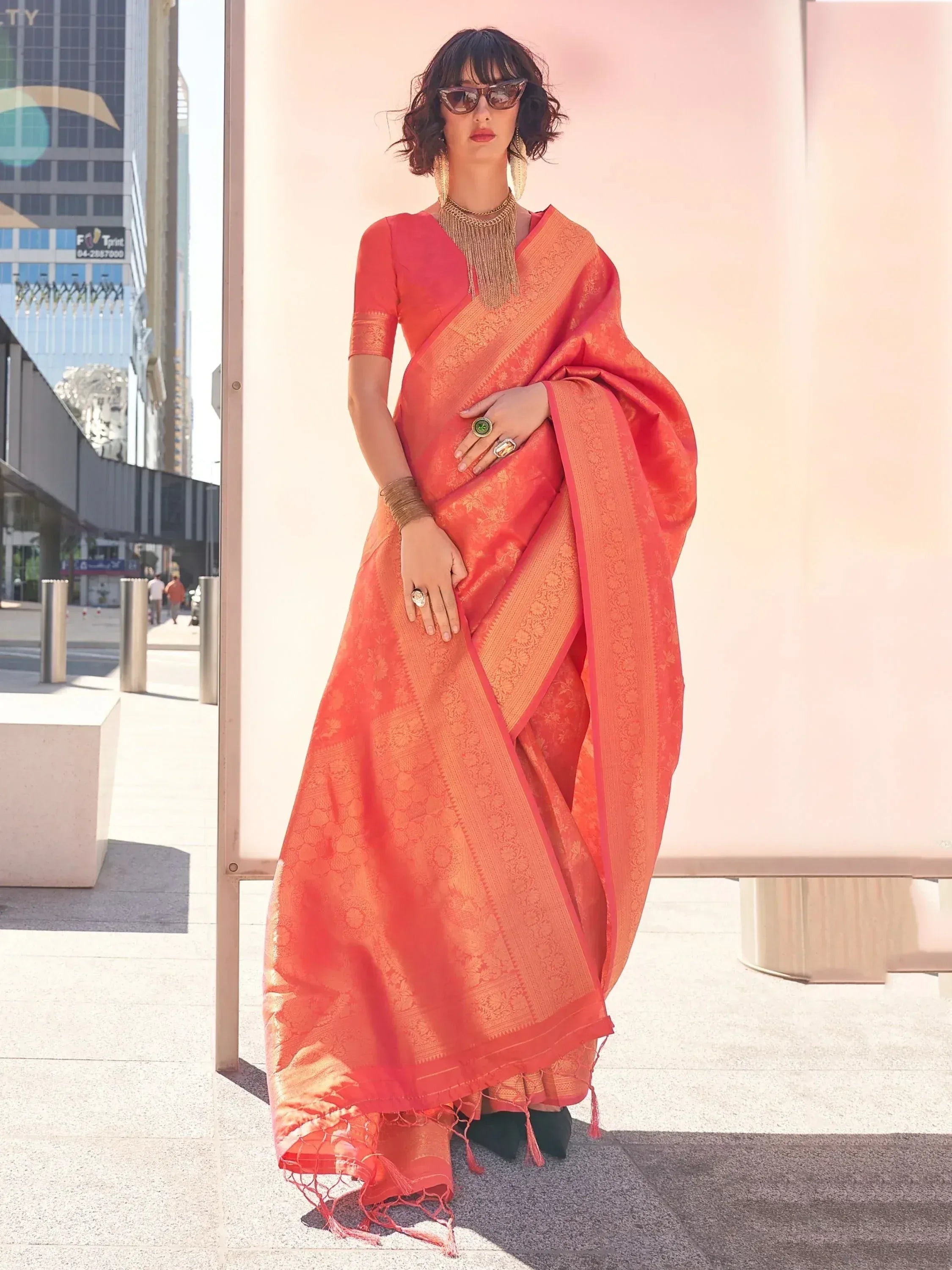 Woman in vibrant coral designer saree with gold zari work, styled with statement jewelry for ethnic wear