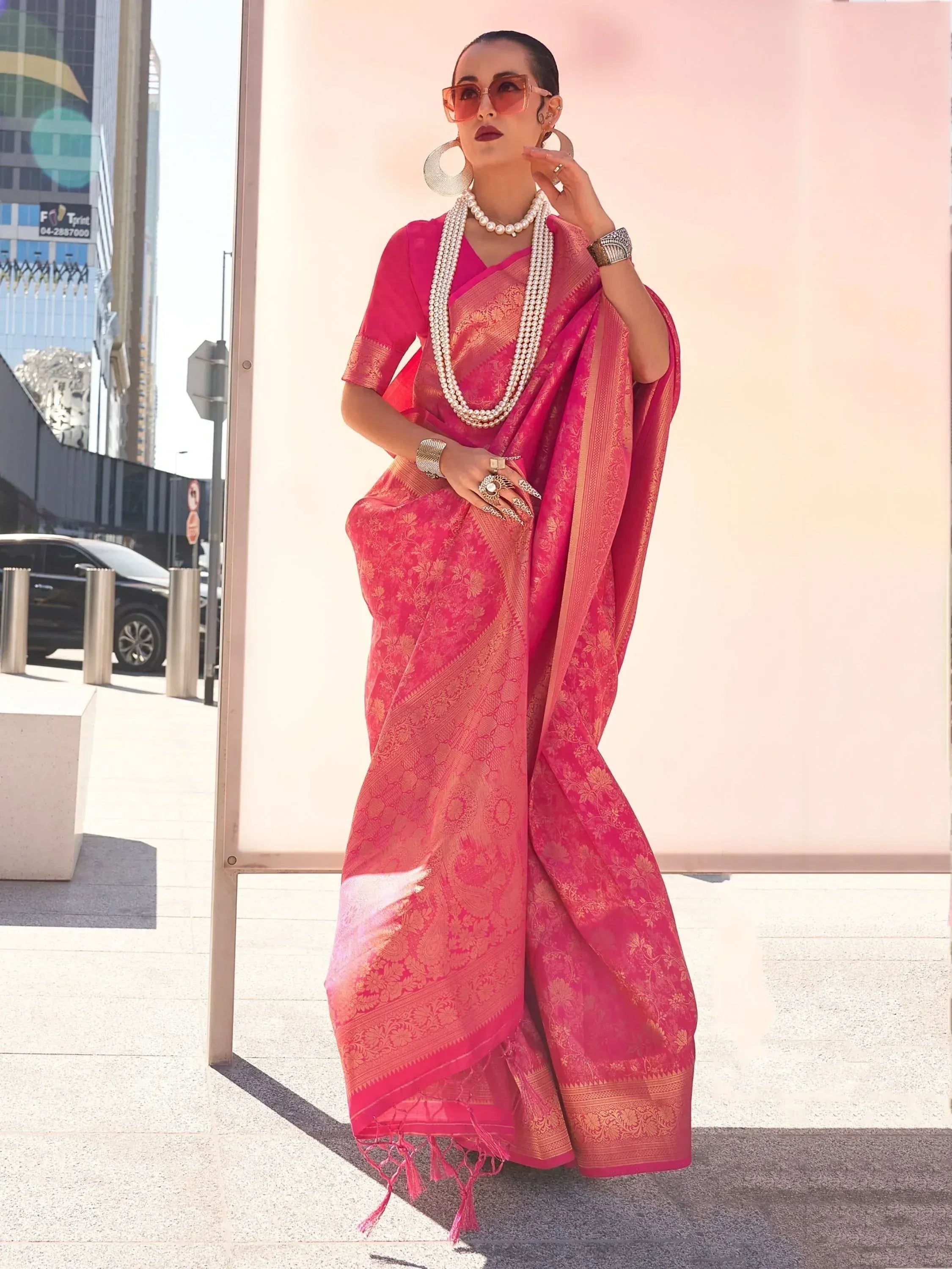Woman in vibrant pink designer saree with gold embroidery, styled with pearl jewelry and sunglasses for modern ethnic look