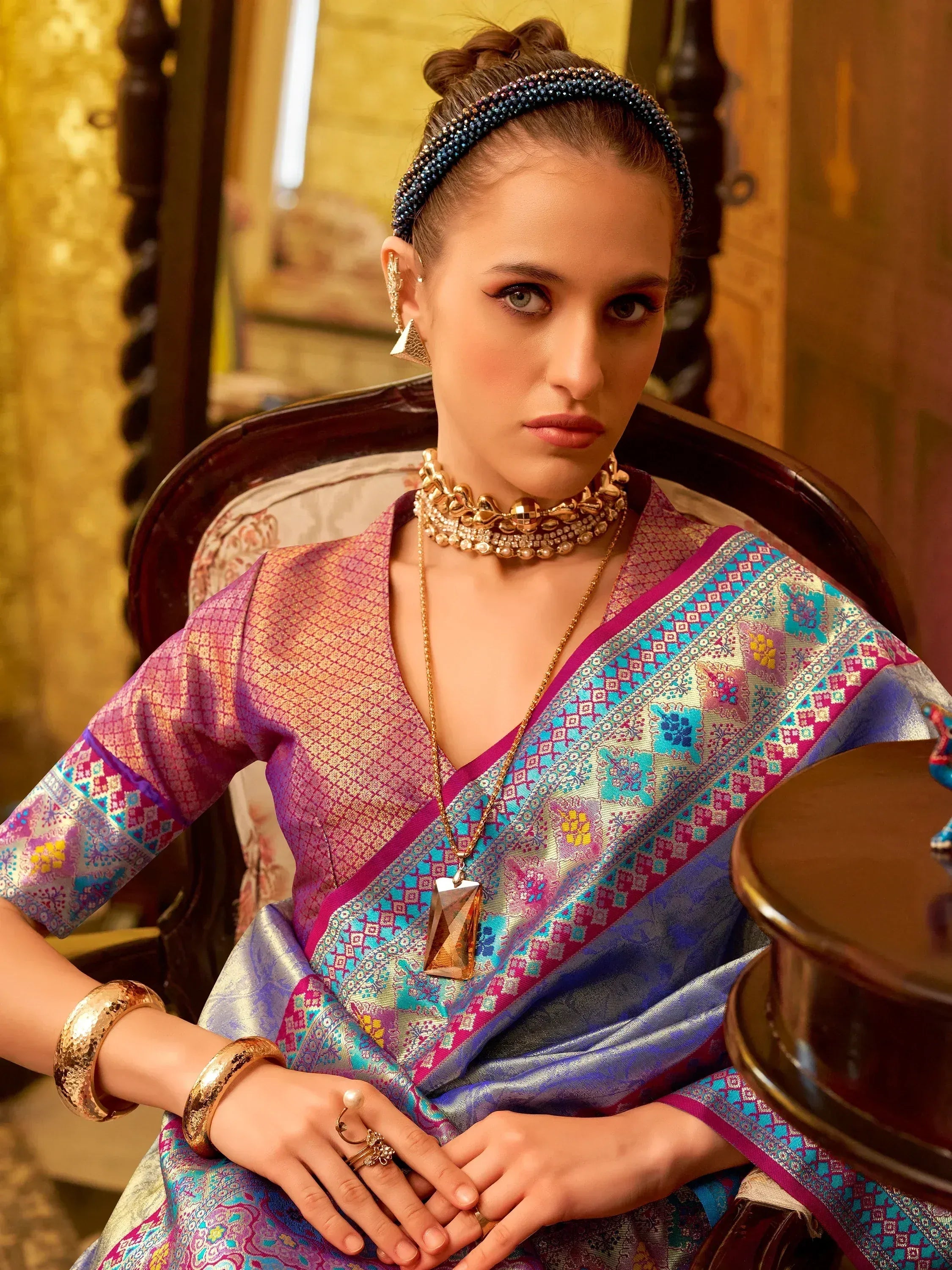 Elegant woman in vibrant Indian saree with intricate patterns and gold jewelry, seated gracefully.