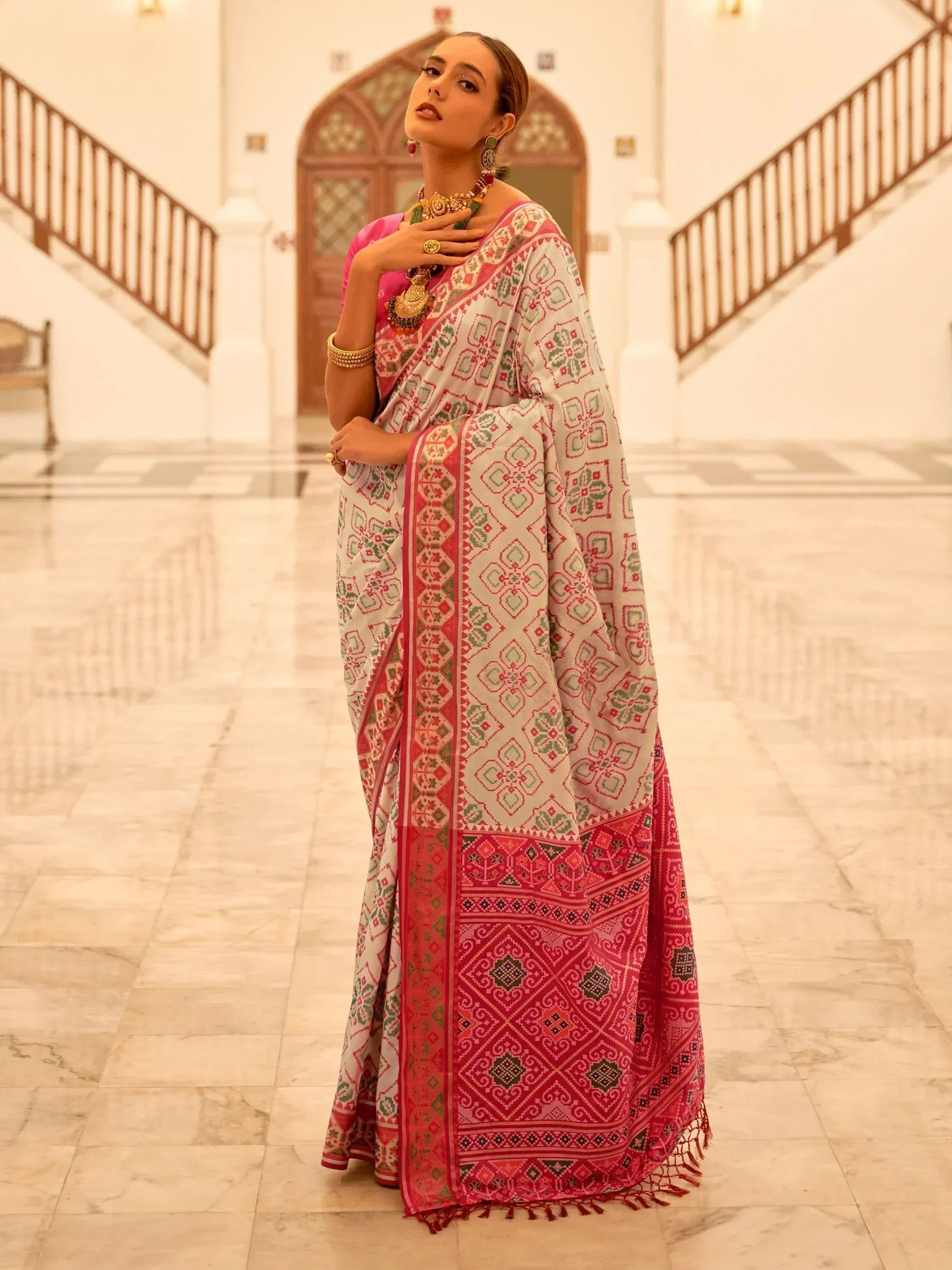 Woman in traditional Indian saree with pink and green geometric print, styled with gold jewelry in palace hallway