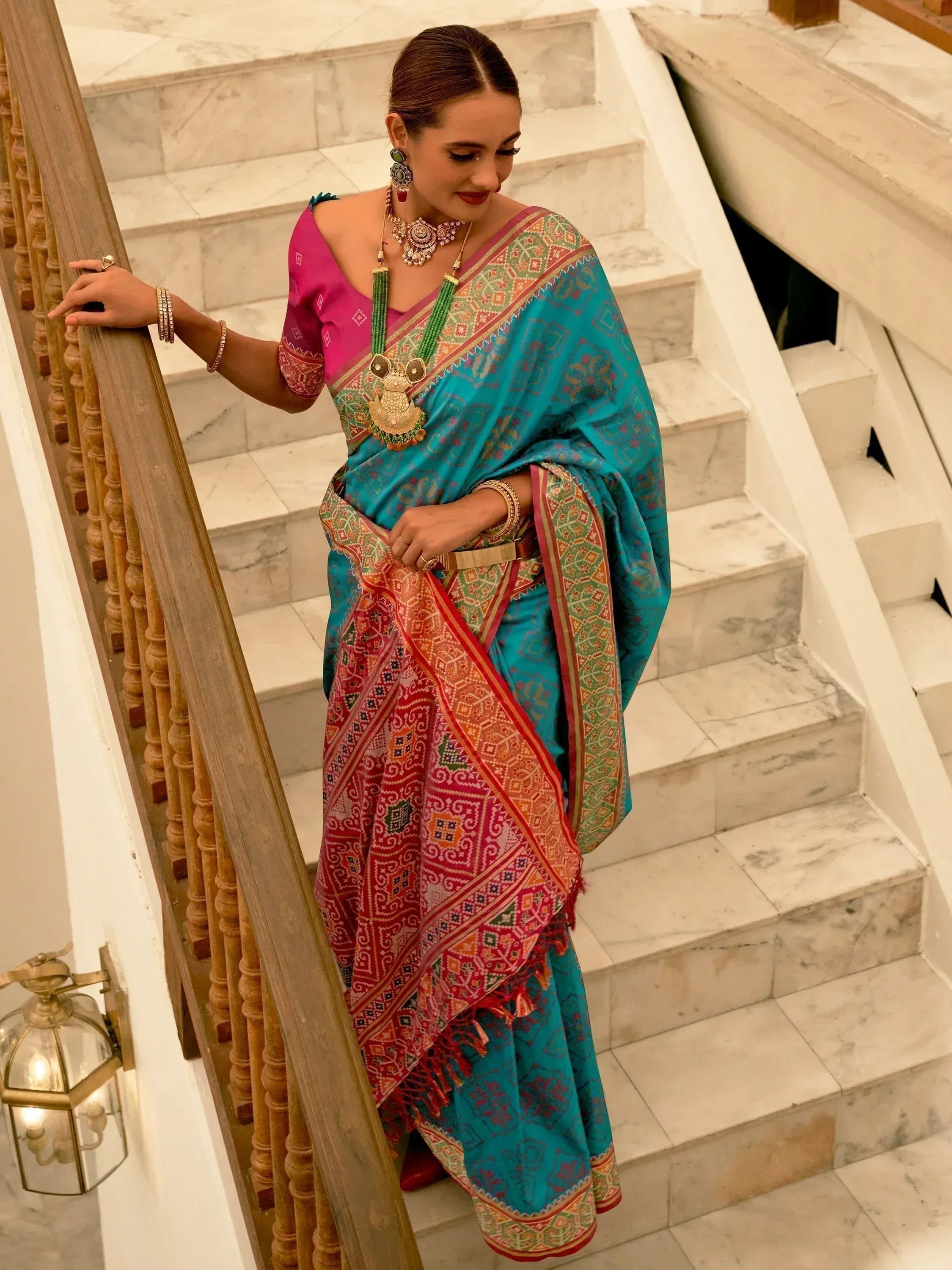 Woman in vibrant turquoise and pink ethnic saree with intricate patterns, standing on marble stairs