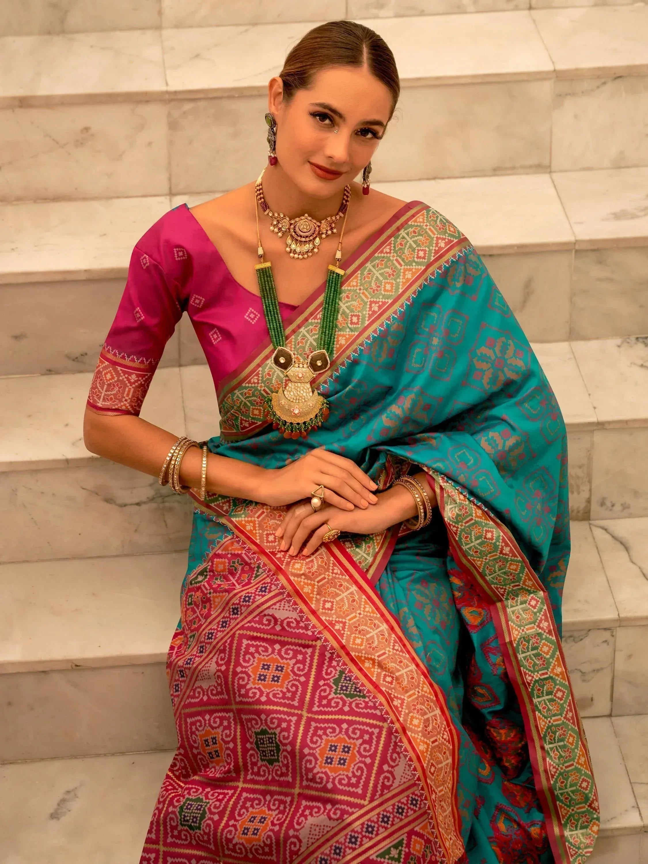 Woman in vibrant turquoise and pink ethnic saree with intricate patterns, traditional jewelry, seated on marble steps