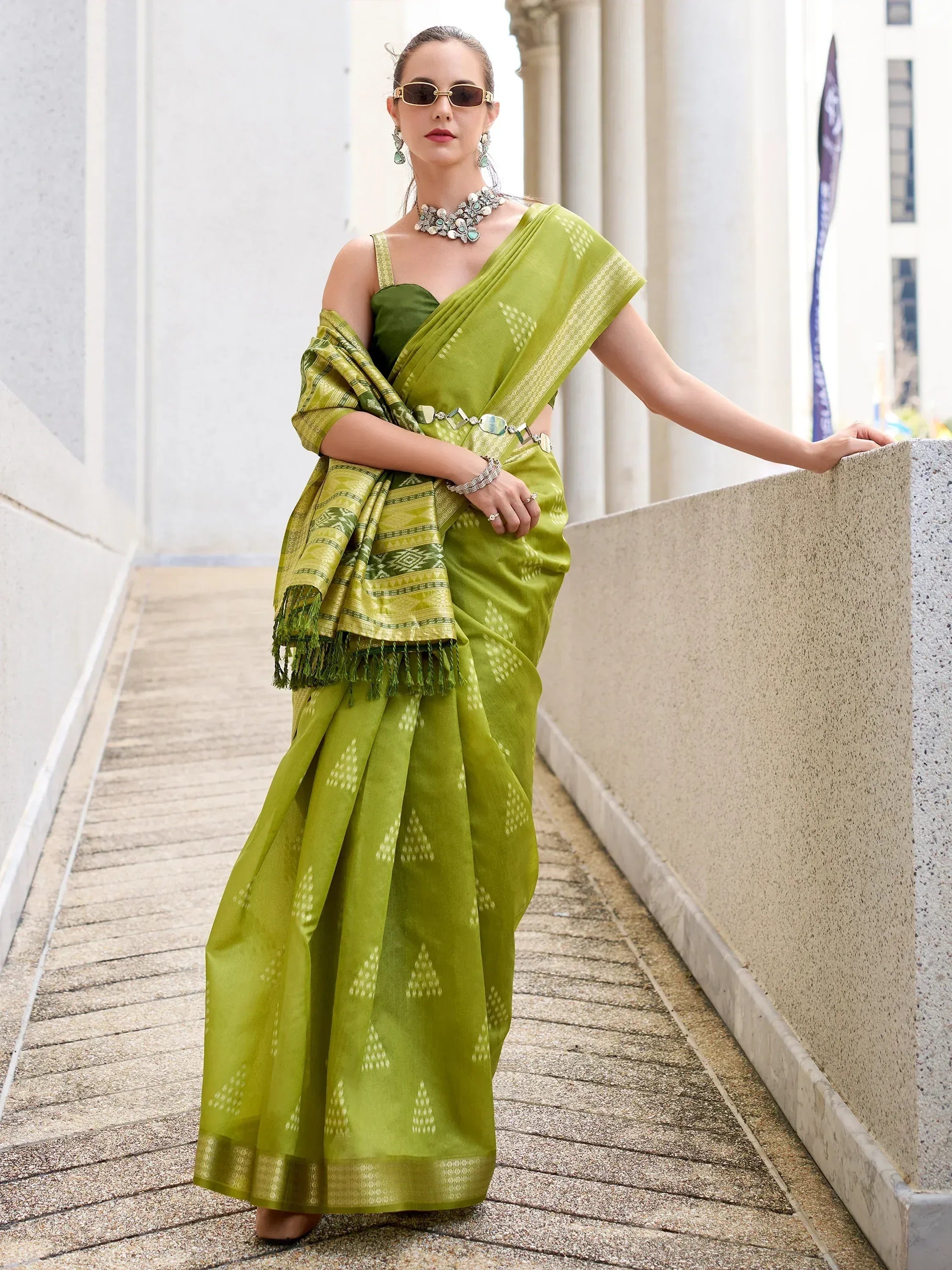 Woman in vibrant green designer saree with geometric patterns, paired with statement jewelry and sunglasses, showcasing ethnic wear.