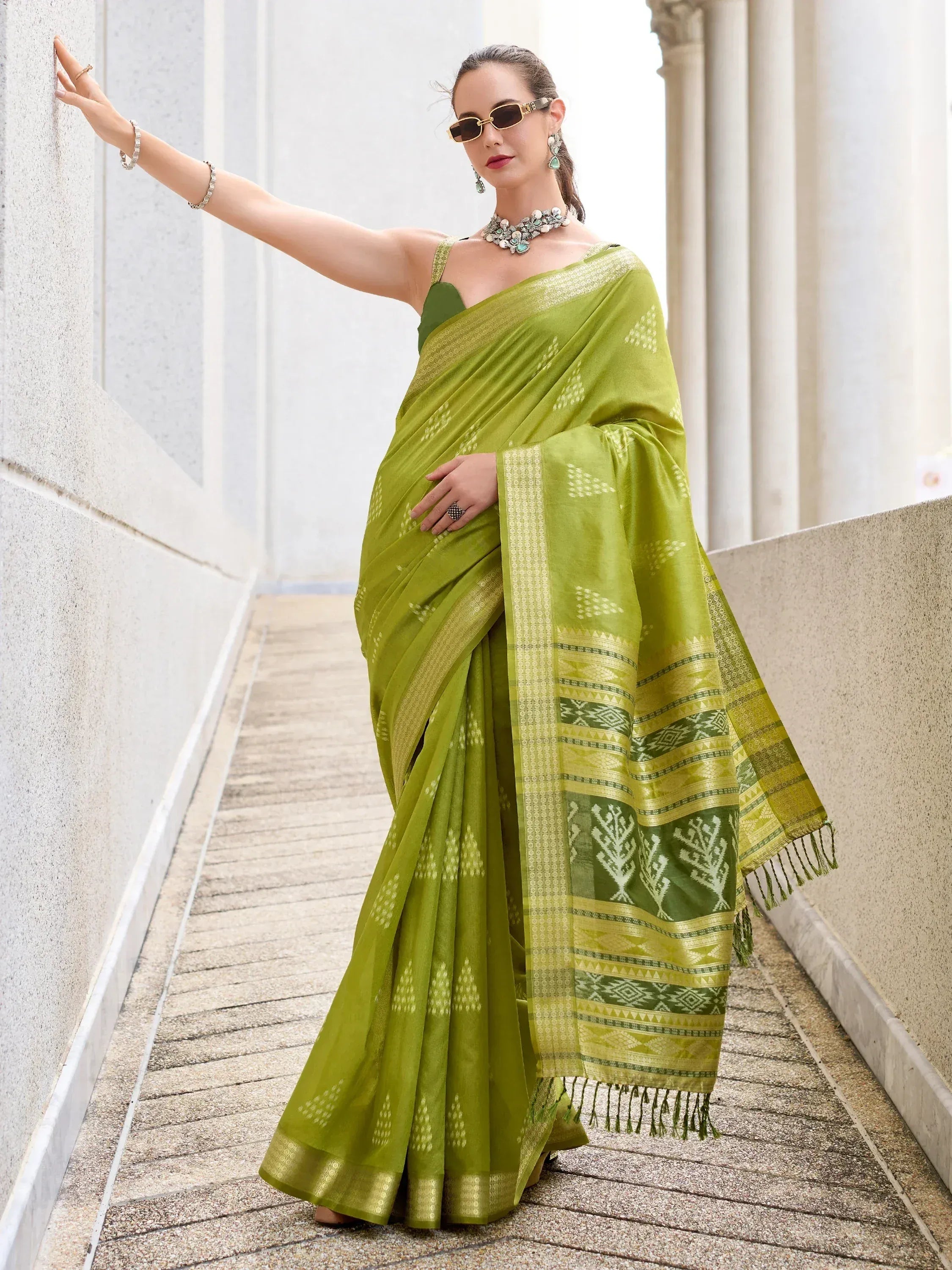 Woman in vibrant green ethnic saree with gold border and geometric patterns, styled with jewelry and sunglasses.