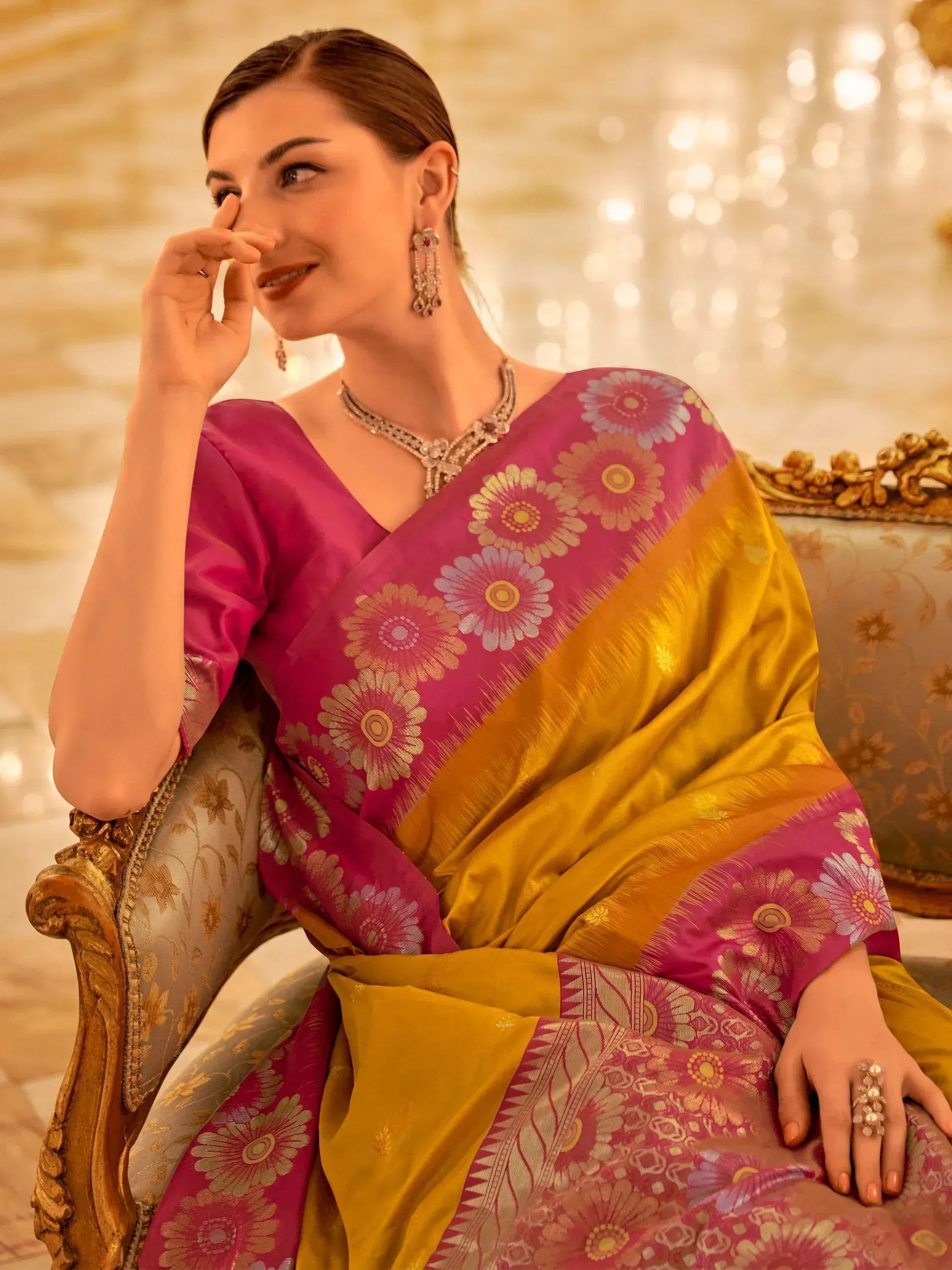 Elegant woman in pink and gold floral saree with traditional jewelry, seated on ornate chair