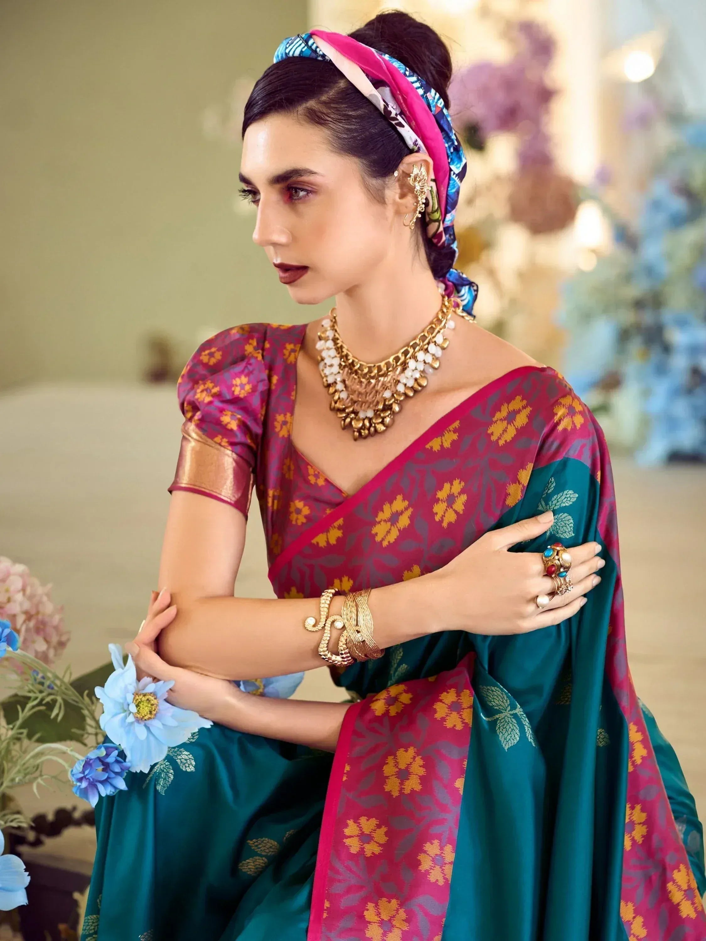 Woman in elegant Indian saree with floral print, gold jewelry, and colorful headscarf, styled for festive occasions.