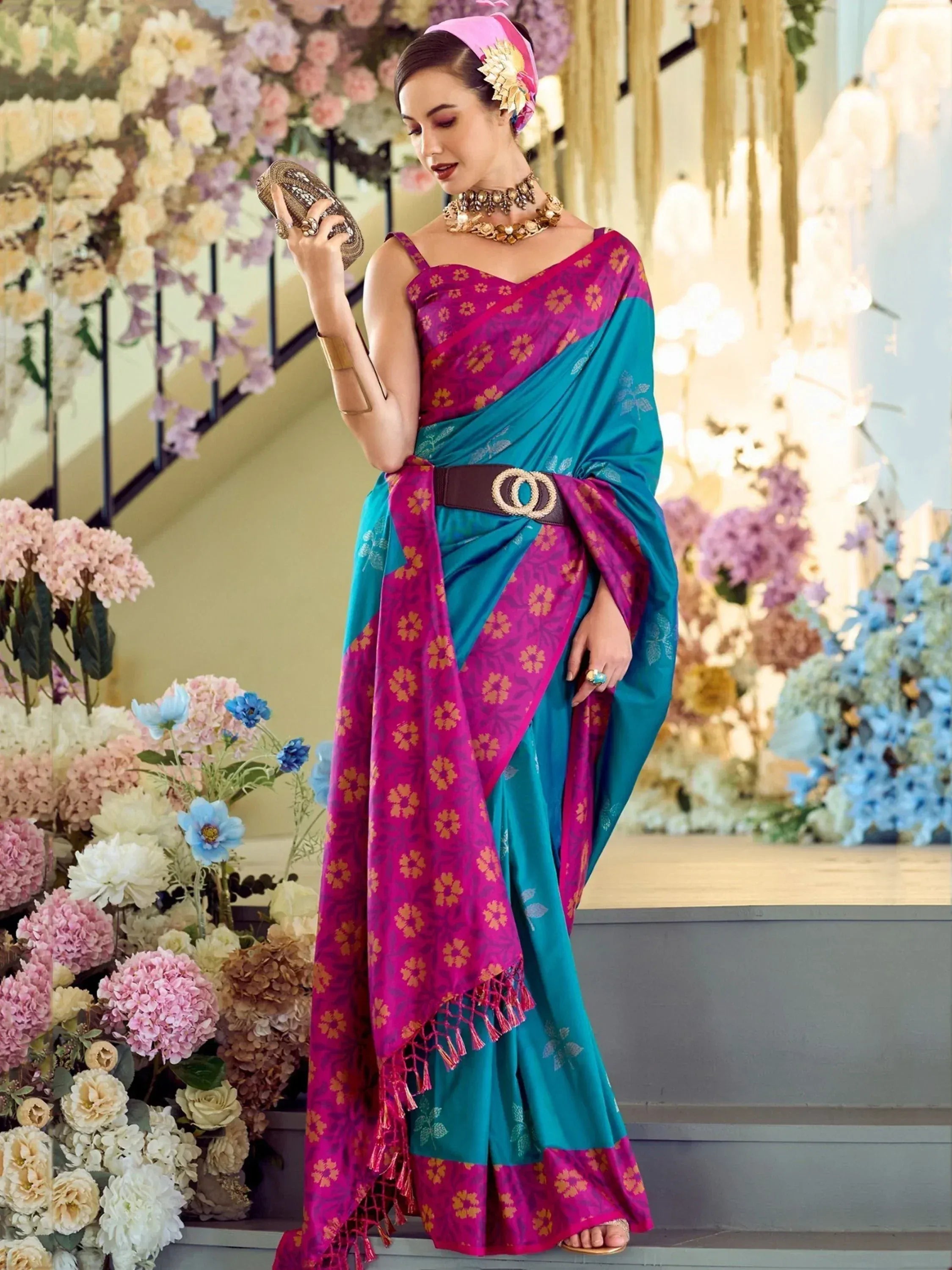 Woman in vibrant pink and teal floral saree with gold jewelry, standing amidst floral decor