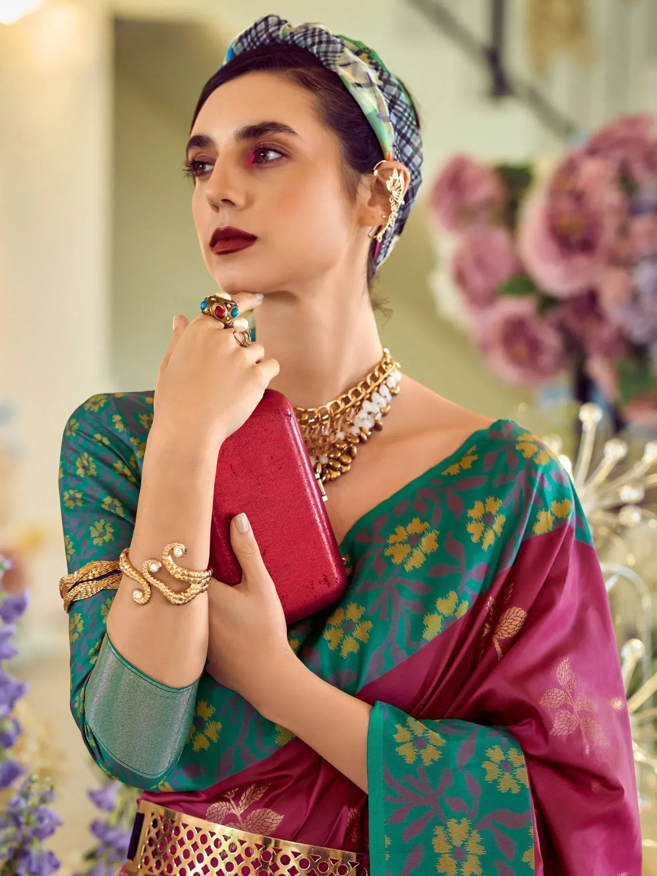 Woman in green and maroon floral saree with gold jewelry and headscarf, holding red clutch