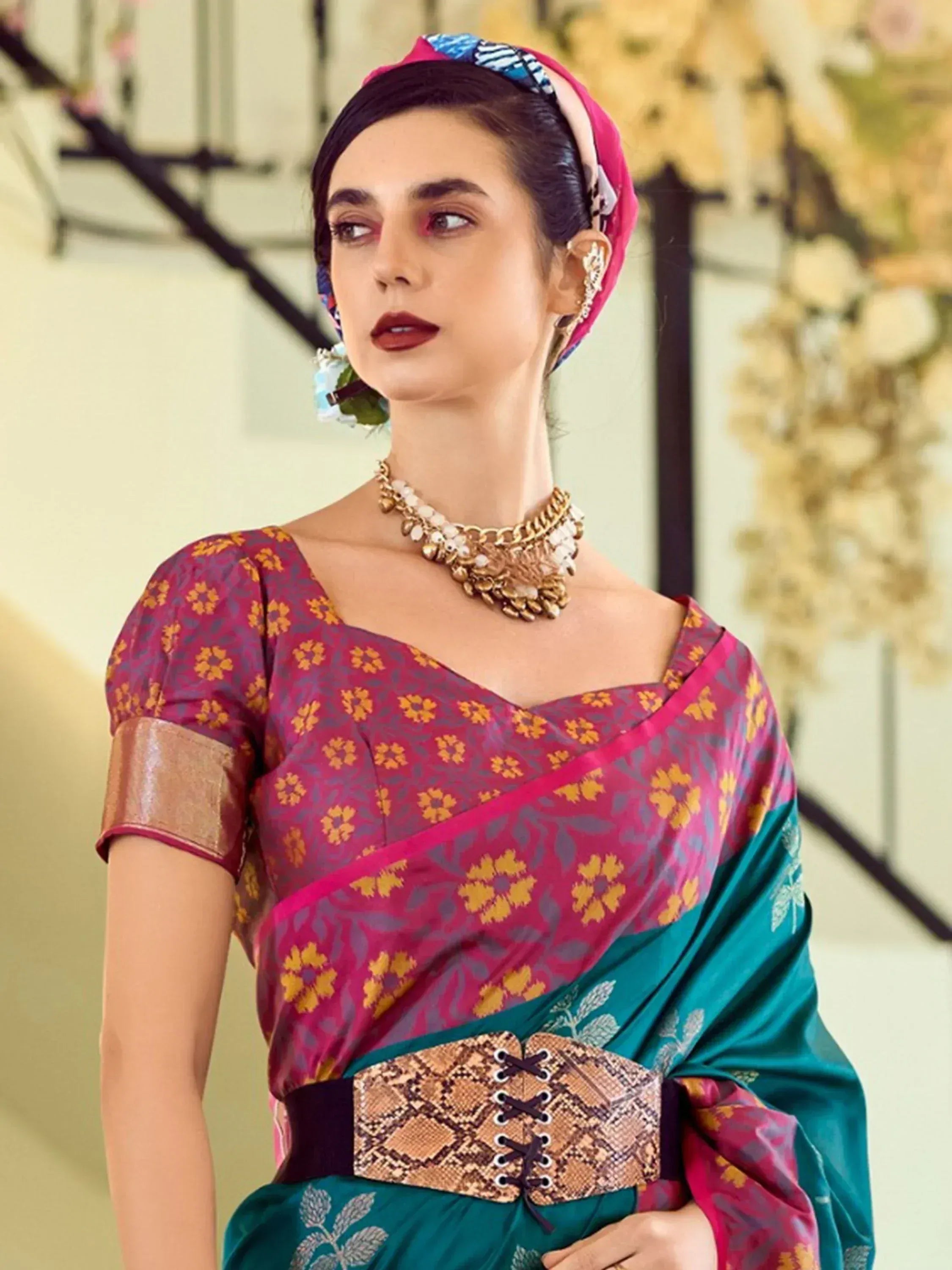 Woman in vibrant floral saree with pink and teal hues, gold jewelry, and patterned headwrap