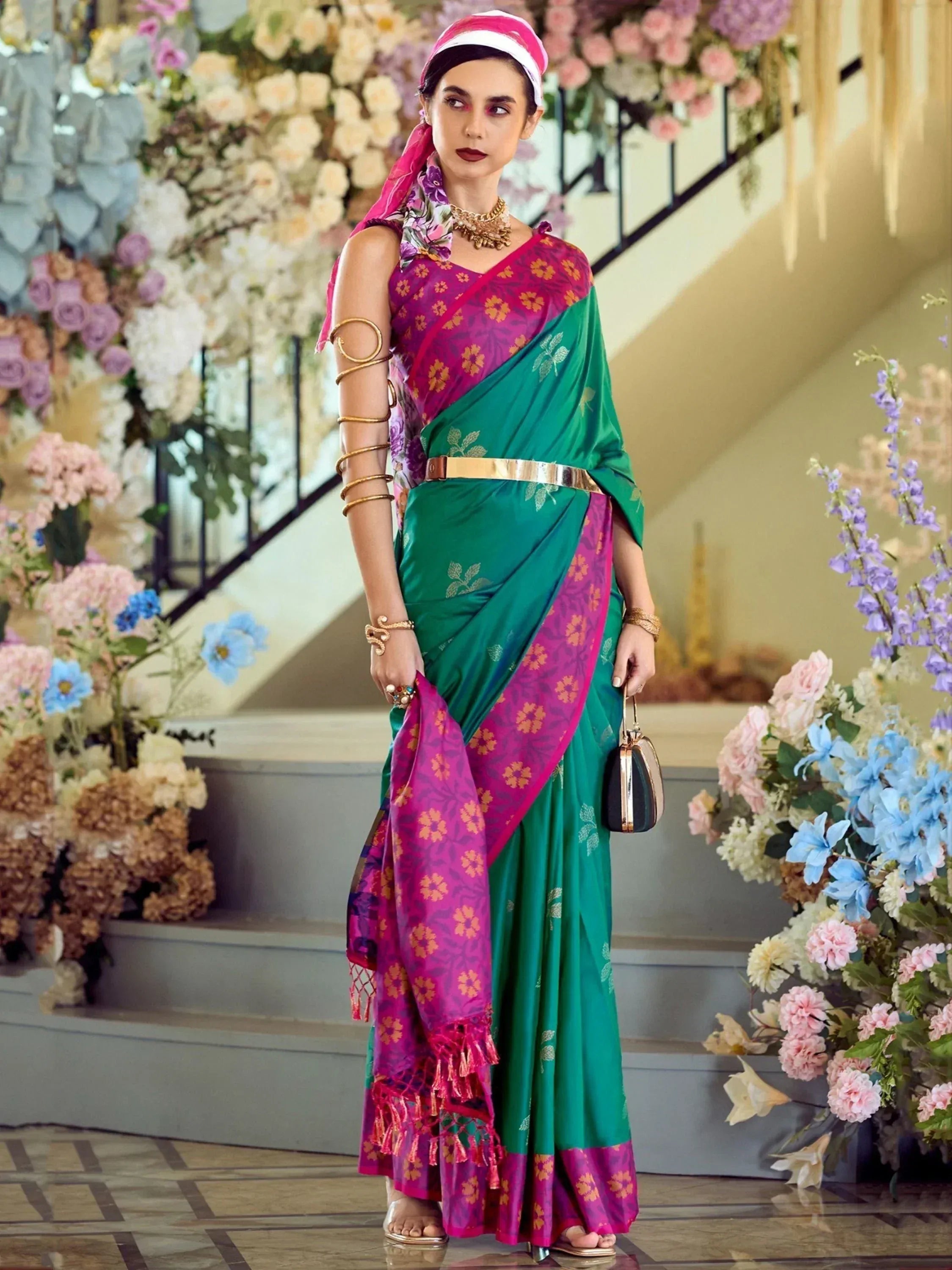 Woman in vibrant green and pink floral saree with gold belt, ethnic jewelry, and floral backdrop