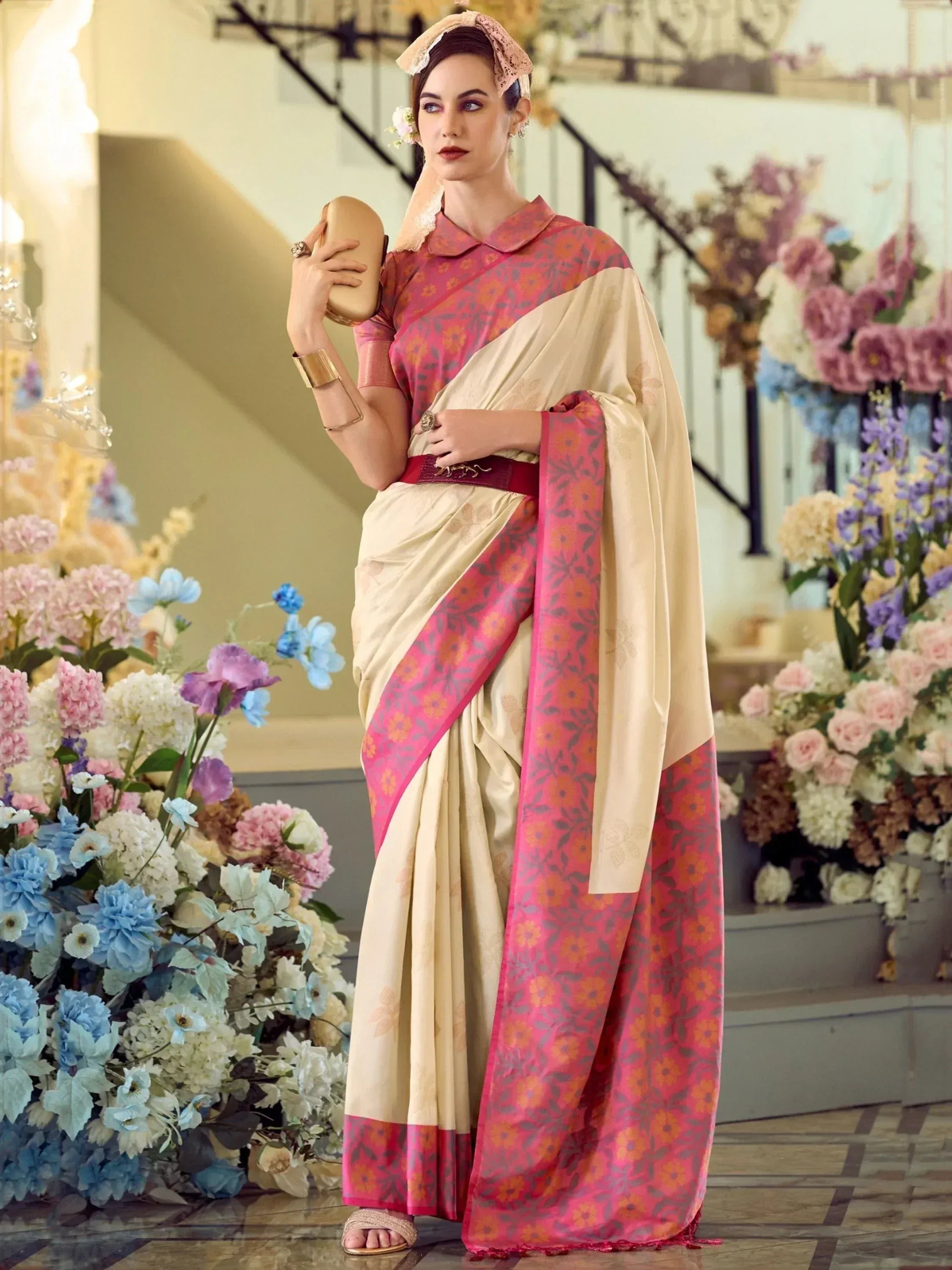 Elegant woman in cream and pink floral saree with lace headpiece, holding clutch, surrounded by flowers