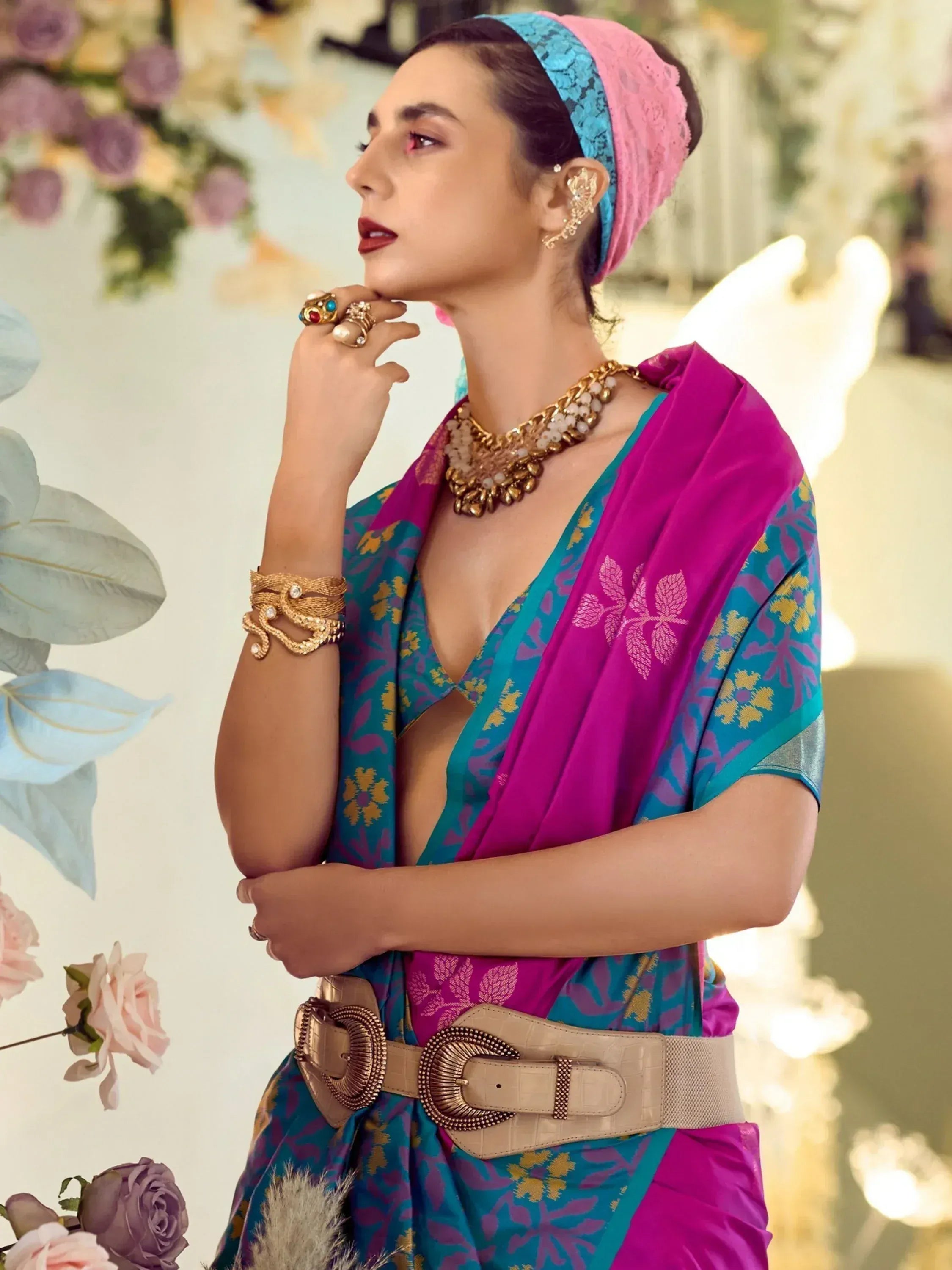 Woman in vibrant blue and pink ethnic saree with gold jewelry, floral backdrop