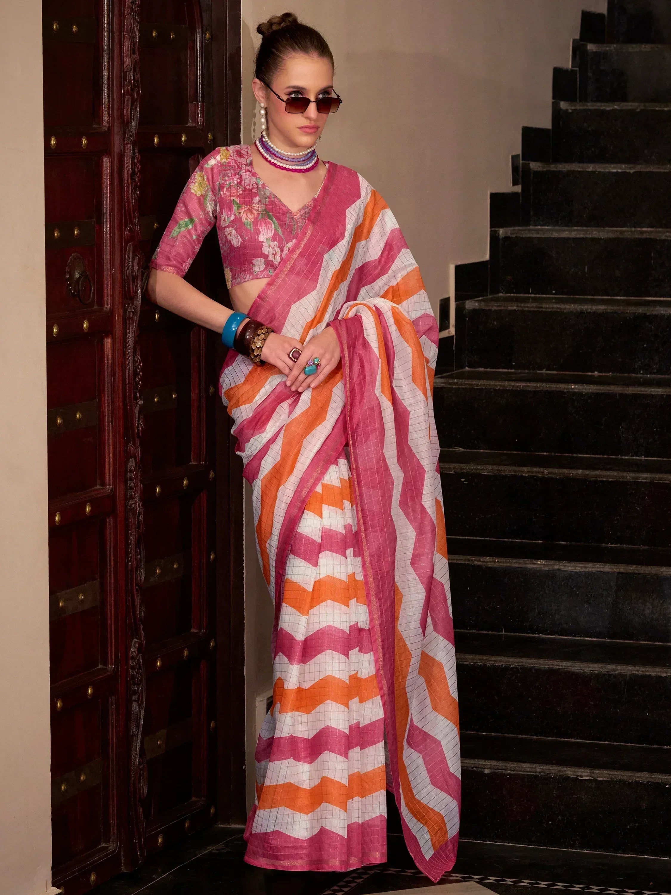 Woman in vibrant pink and orange chevron saree with floral blouse, styled with sunglasses and jewelry.