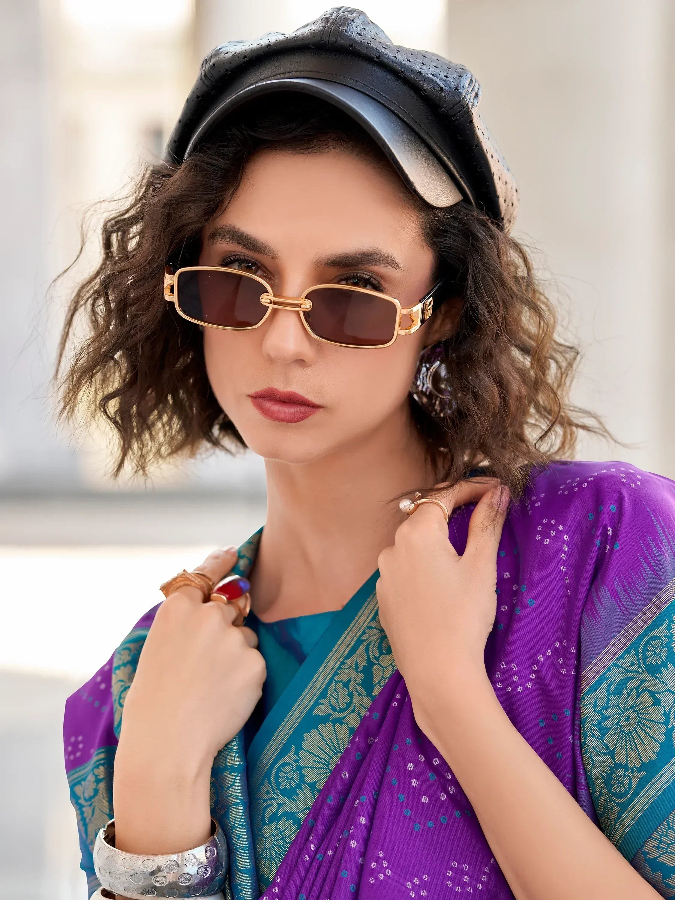 Woman in purple and teal saree with gold sunglasses and black cap, styled for ethnic fashion