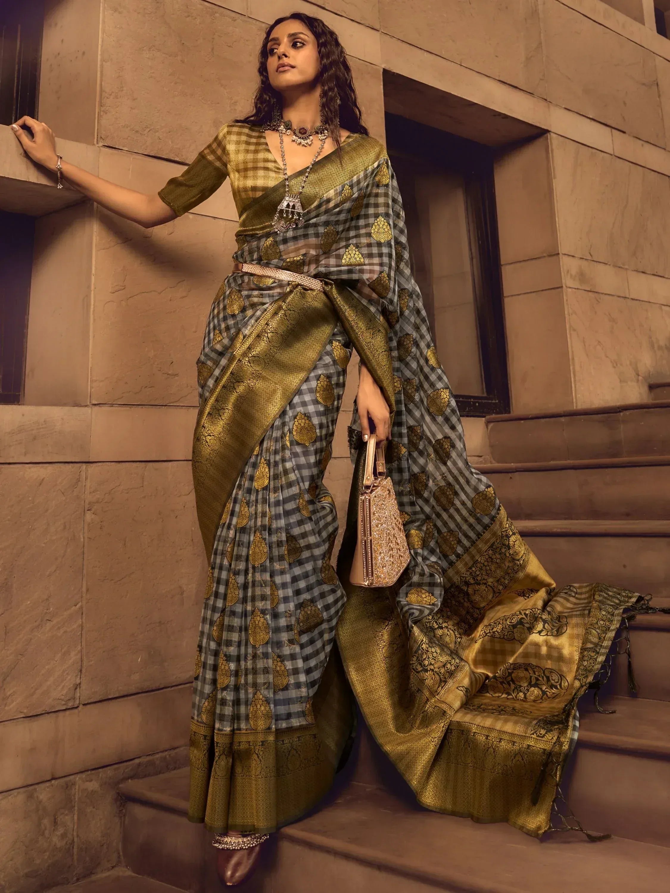 Woman in elegant grey and gold checkered saree with intricate golden motifs, styled with traditional jewelry and clutch.
