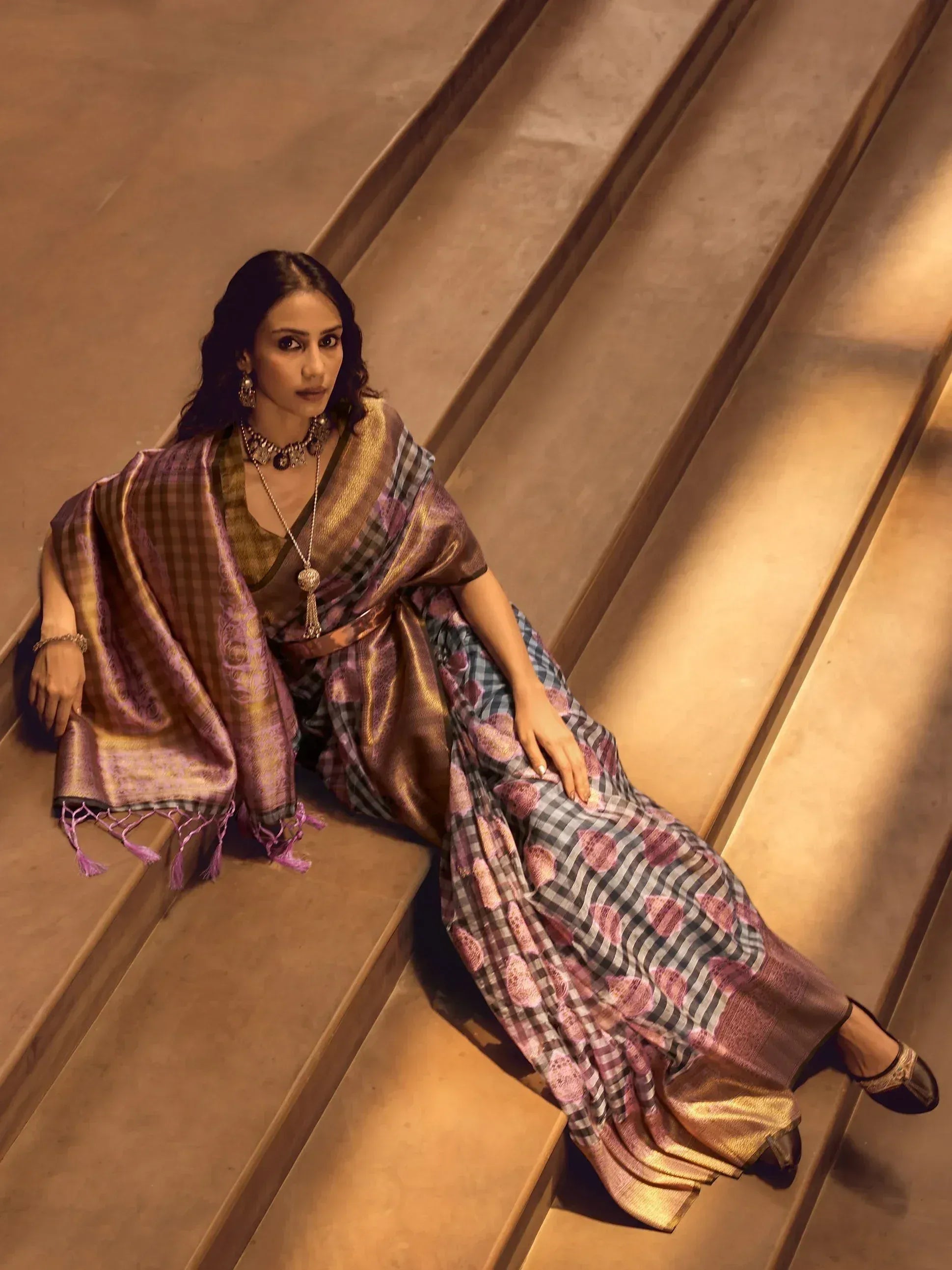 Woman in purple and gold checkered saree with floral motifs, styled with ethnic jewelry, seated on stairs.